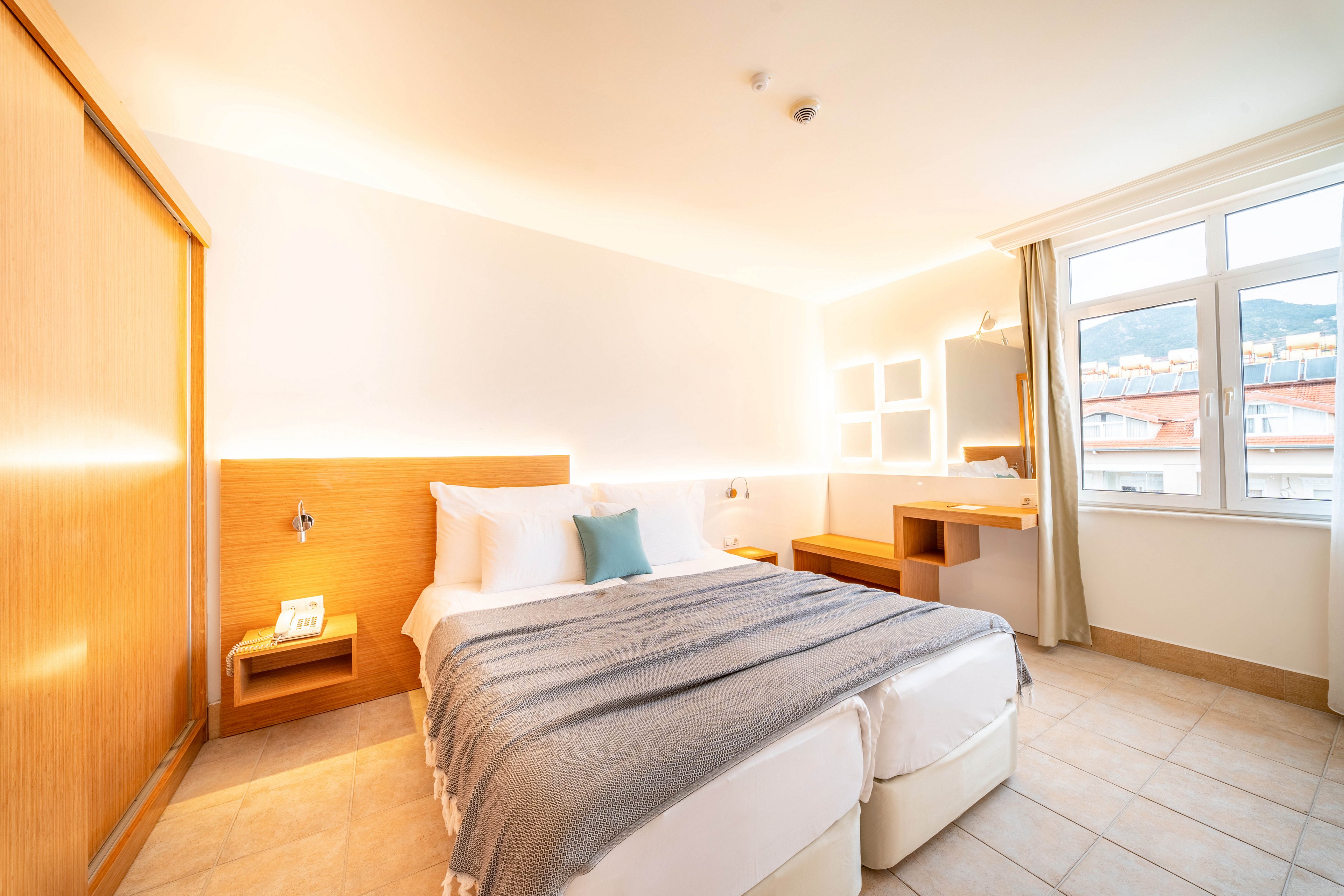 A contemporary hotel bedroom features a neatly made double bed with white linens and a gray throw blanket. Warm wooden furniture, including a bedside shelf and desk, contrasts with the light tiled floor and white walls. Large windows let in abundant natural light and reveal a view of nearby buildings and hills. Soft ambient lighting around the headboard and mirrors creates a cozy, inviting atmosphere.