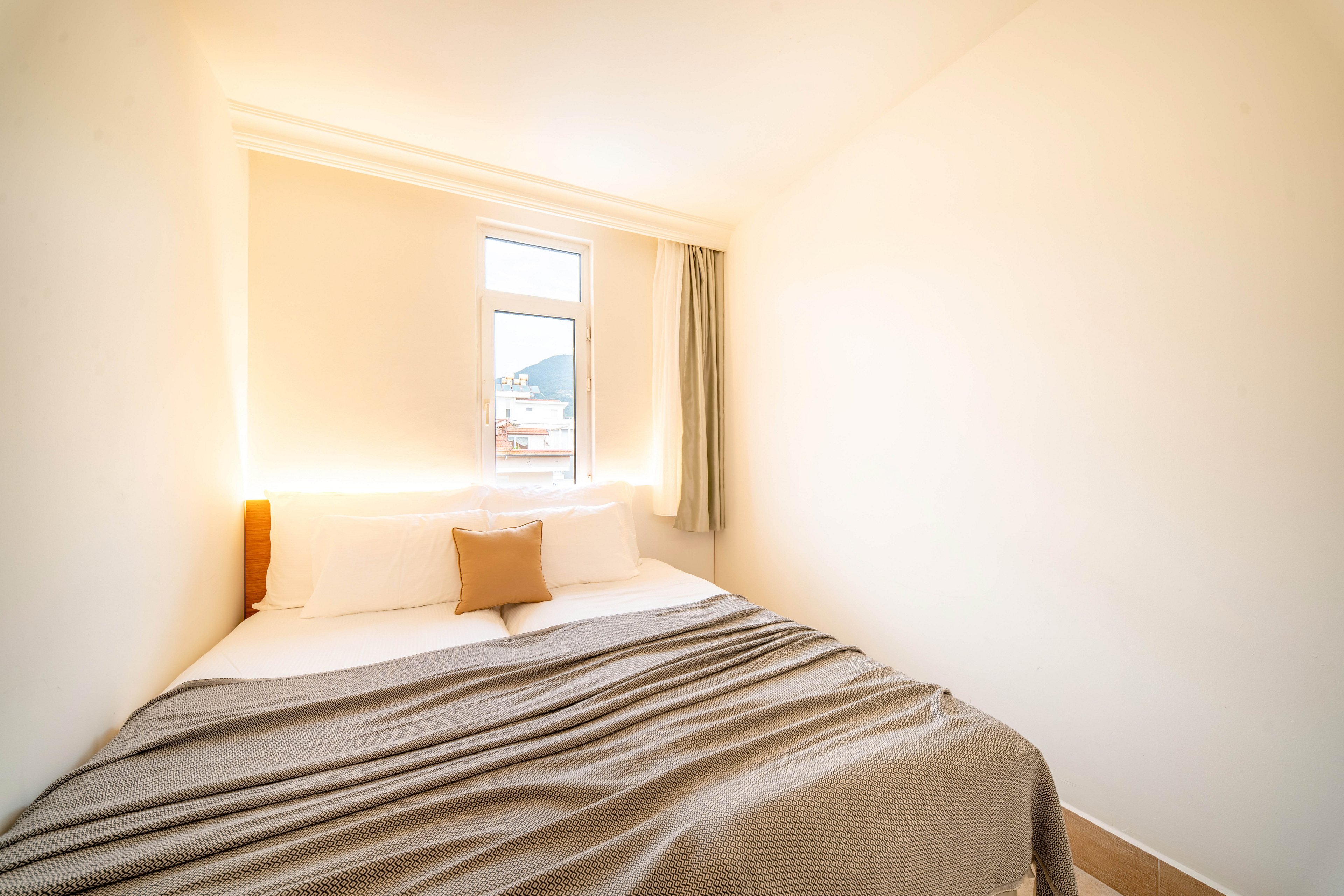 A small, bright bedroom features a neatly made double bed with white pillows and a single beige accent cushion. Soft natural light streams in through a narrow window at the end of the room, illuminating the simple cream-colored walls. A textured gray blanket is spread smoothly over the bed, adding subtle contrast to the light decor. A curtain partially covers the window, framing a glimpse of the outdoors and enhancing the serene, minimalist atmosphere.