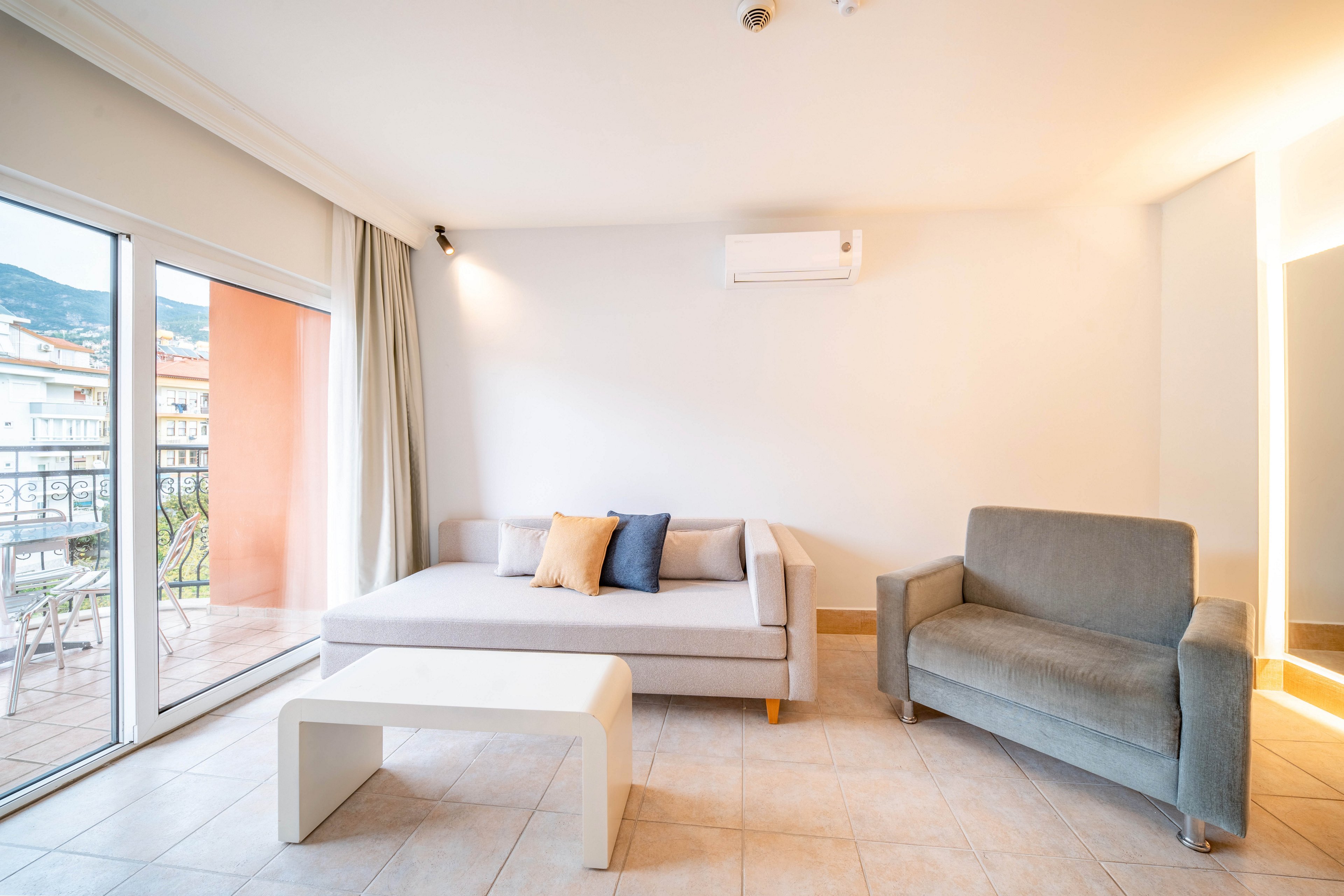 A contemporary living room features a light gray sofa with colorful accent pillows and a matching armchair. Large sliding glass doors open to a balcony, allowing natural light to fill the space and reveal an exterior cityscape with hills in the background. The room has neutral tiled flooring, white walls, and a minimalist white coffee table. An air conditioning unit is mounted high on the wall, contributing to the clean, modern aesthetic.