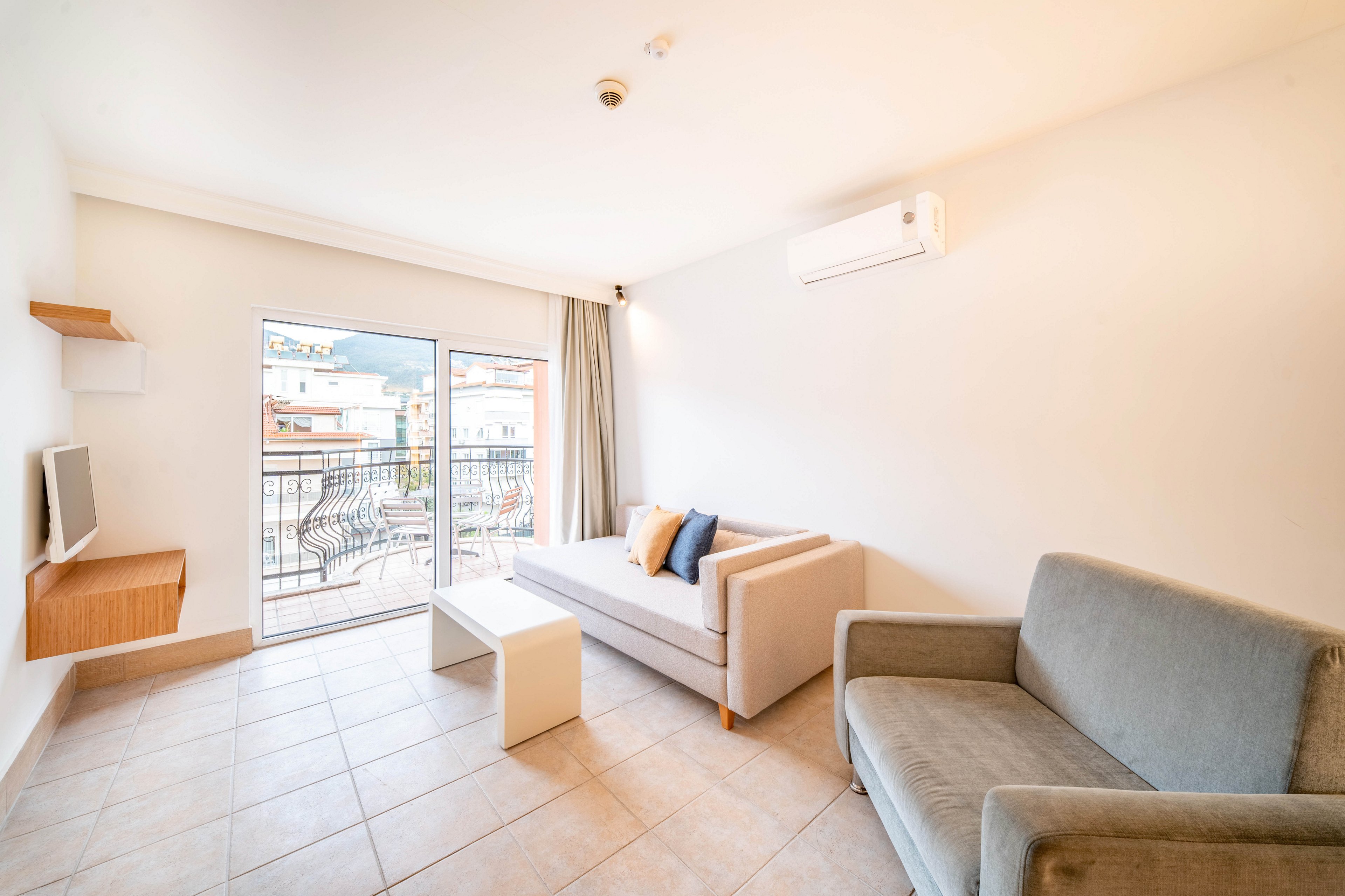 A minimalist living room features a light-colored sofa and an armchair arranged around a small white coffee table. Large glass doors open to a balcony with outdoor furniture and a view of neighboring buildings and hills. The space is decorated in neutral tones with tiled flooring, a wall-mounted television, and a compact air conditioning unit. Natural daylight fills the room, creating an airy and inviting atmosphere.