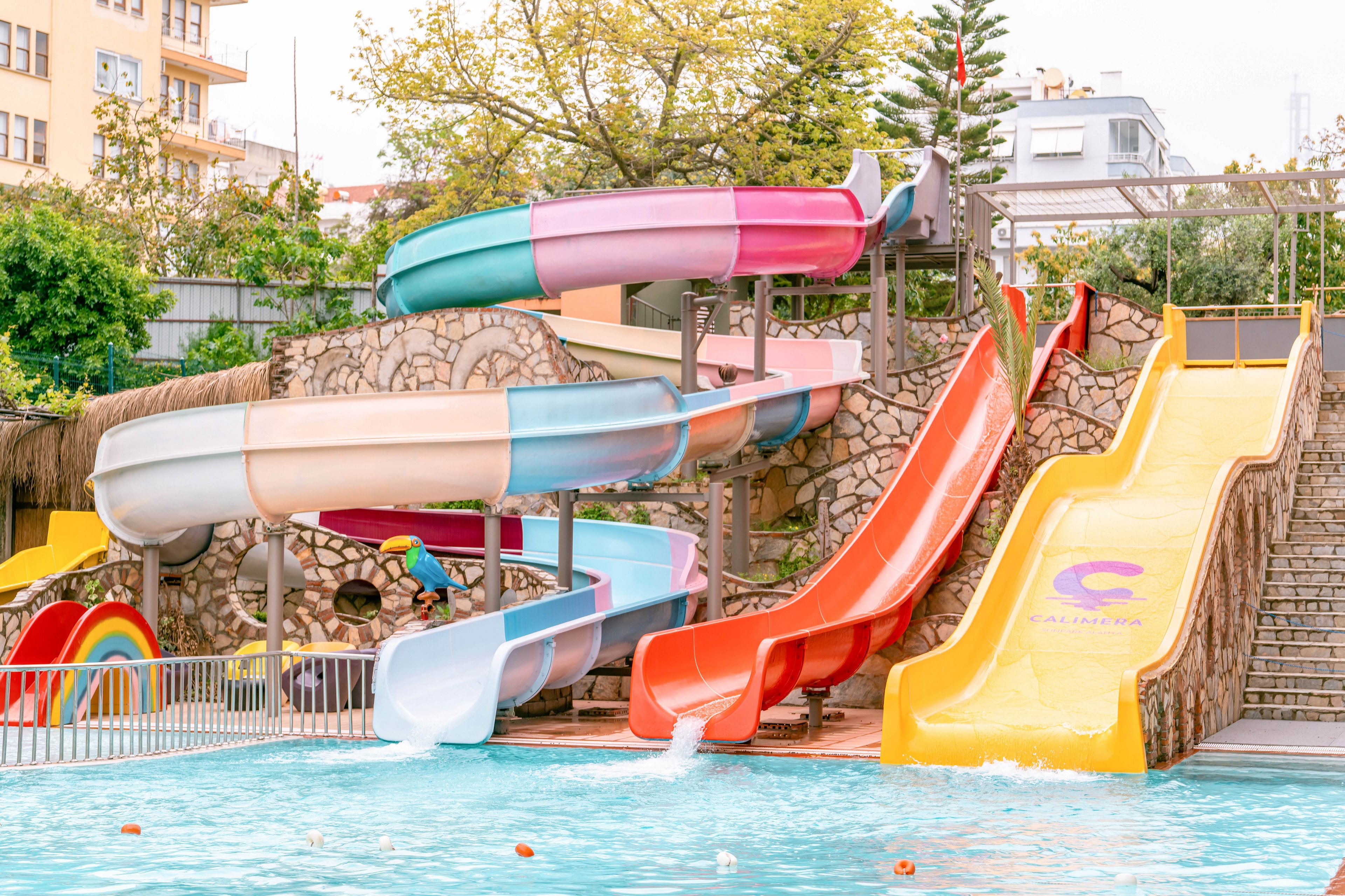 A group of bright, curving water slides descends into a clear blue swimming pool. The slides are painted in pastel and vivid colors, including pink, blue, orange, yellow, and white. Stone walls and railings frame the attraction, with trees and nearby buildings visible in the background. The scene suggests a fun, family-friendly outdoor water park on a sunny day.