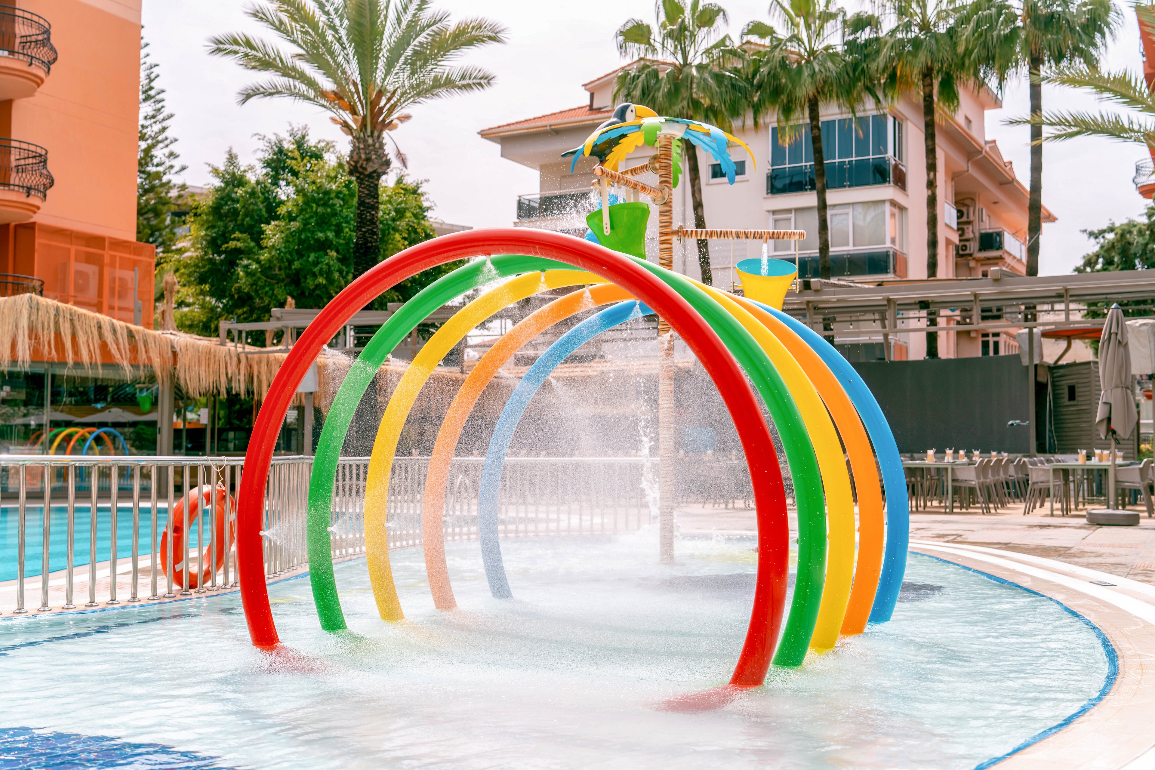 A series of brightly colored rainbow arches spray water over a shallow resort pool. The splash pad structure is set outdoors near palm trees and multi-story hotel buildings. Clear blue water and mist from the fountains create a refreshing, playful atmosphere. Lounge chairs and railings in the background suggest a family-friendly vacation setting.