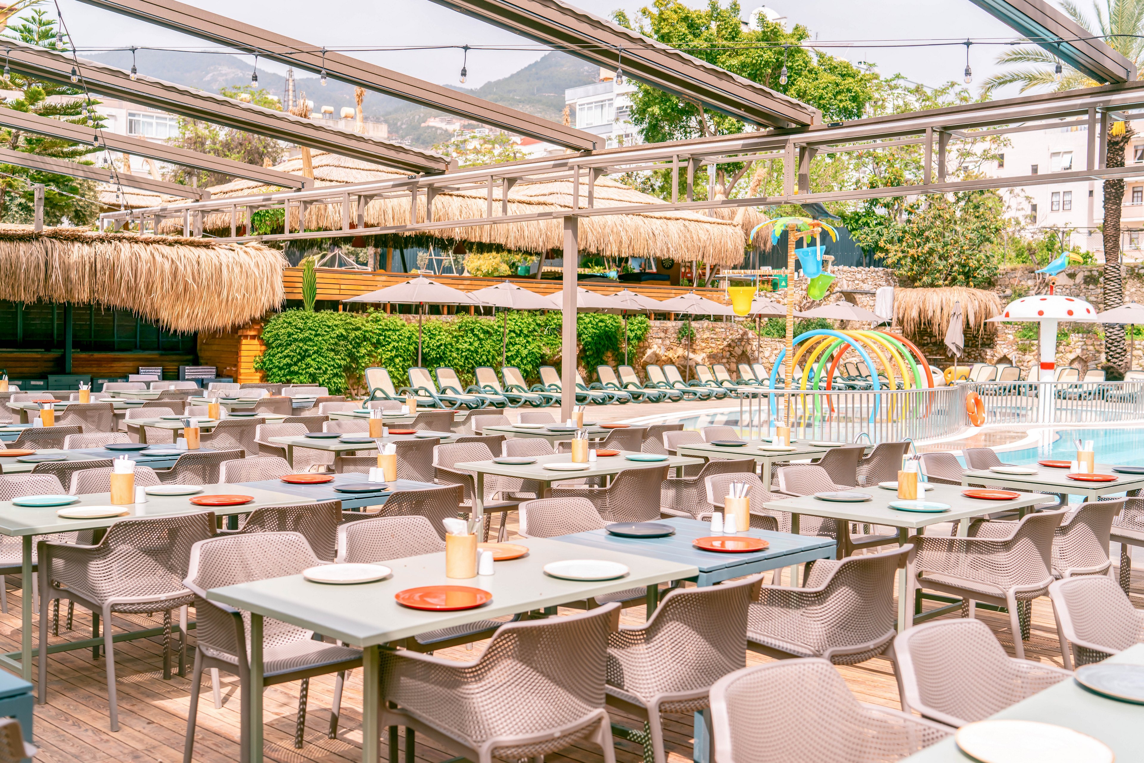 Bright outdoor dining area with rows of tables and chairs is set beside a colorful resort swimming pool. Plates, cups, and condiments are neatly arranged on the tables, suggesting readiness for guests. A thatched roof bar and sun loungers line the background, with playful water arches and slides in the pool area. Overhead metal frames and string lights add structure and ambiance, while lush greenery and distant buildings frame the scene.