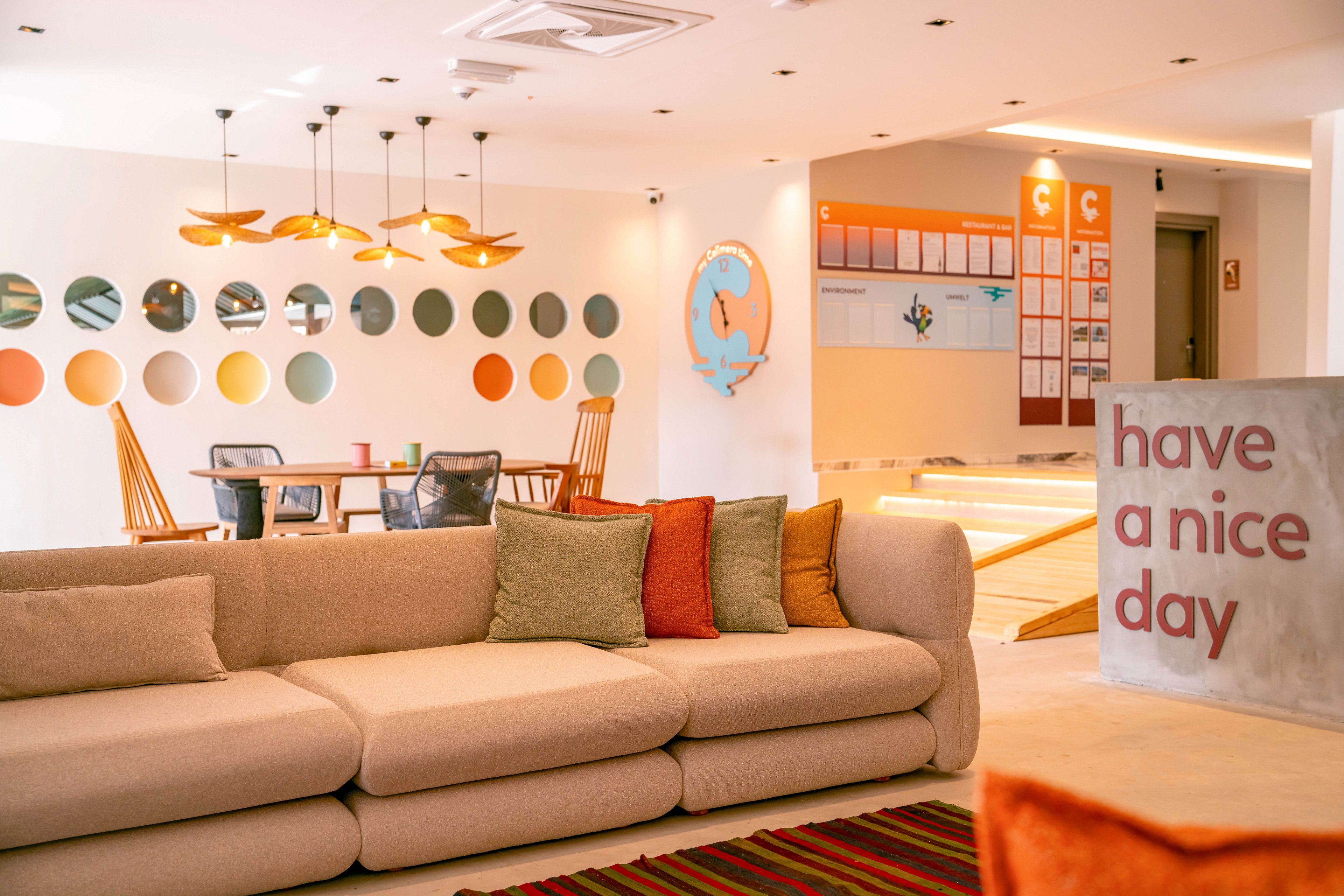 Bright contemporary lounge area featuring a large beige sofa with colorful cushions. The setting includes a dining table with wooden chairs and a wall decorated with circular color swatches. Warm pendant lights hang from the ceiling, and a striped rug adds texture to the floor. A concrete pillar in the foreground displays the phrase "have a nice day," enhancing the welcoming atmosphere.