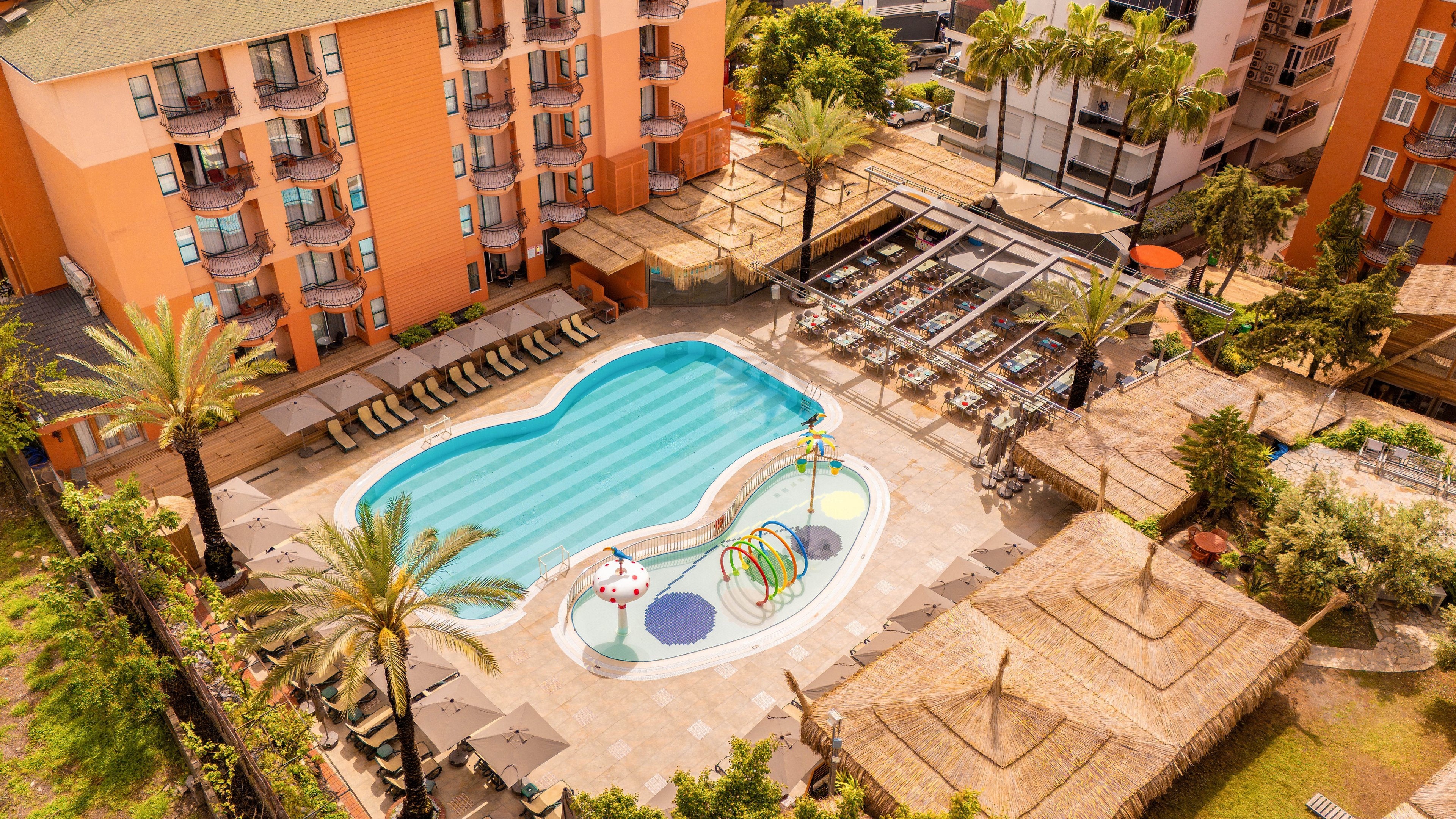 A resort-style outdoor swimming pool is seen from above, surrounded by sun loungers and palm trees. The pool area includes a colorful children's splash zone with water features and shaded seating nearby. Adjacent buildings with balconies and orange facades frame the courtyard, while cabanas and covered terraces provide additional relaxation spaces. The warm lighting and tidy layout suggest a sunny vacation destination focused on leisure and family fun.