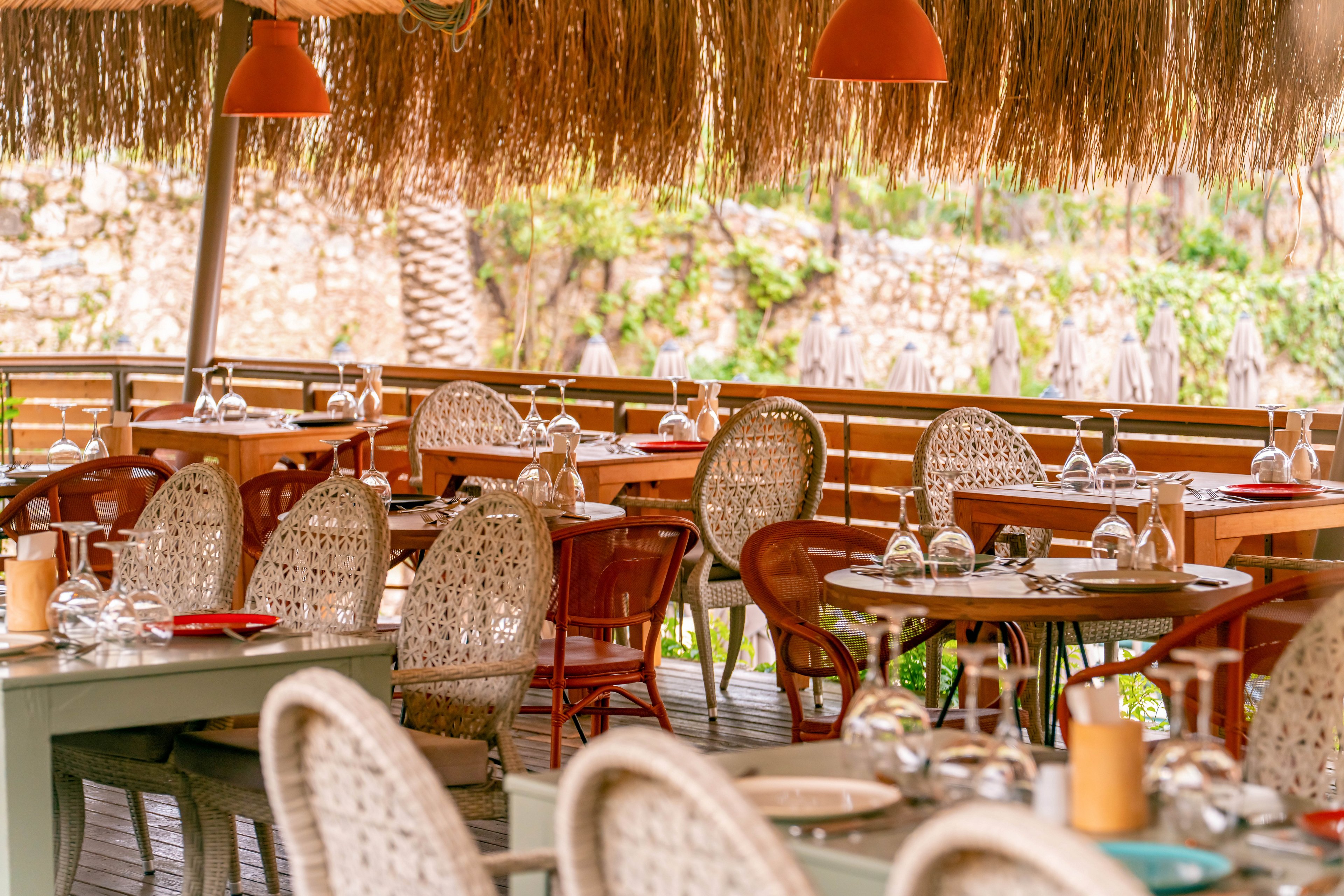 An open-air restaurant terrace features neatly arranged tables ready for guests. Wicker chairs surround round wooden tables set with plates, cutlery, and upside-down wine glasses. Warm orange pendant lights hang from a thatched roof, creating a cozy atmosphere. The background shows greenery and stone walls, adding a natural, relaxed setting.