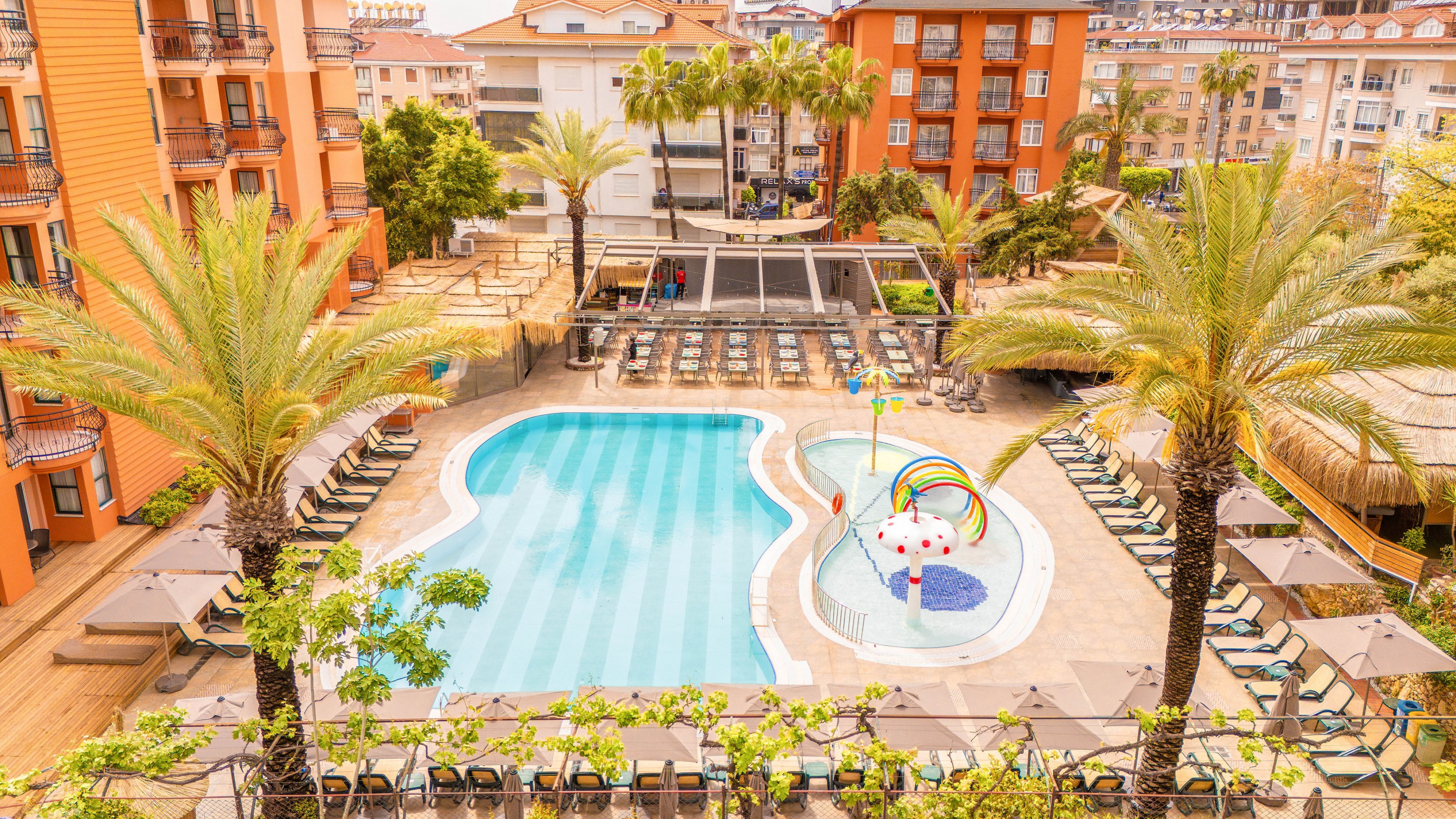 An elevated view shows a large resort swimming pool surrounded by lounge chairs and tall palm trees. The pool area features a shallow section with colorful water play structures, including a rainbow arch and playful figures. Warm-toned buildings with balconies frame the courtyard, while additional residential structures are visible in the background. The bright daylight and clear water create a relaxed, vacation atmosphere.