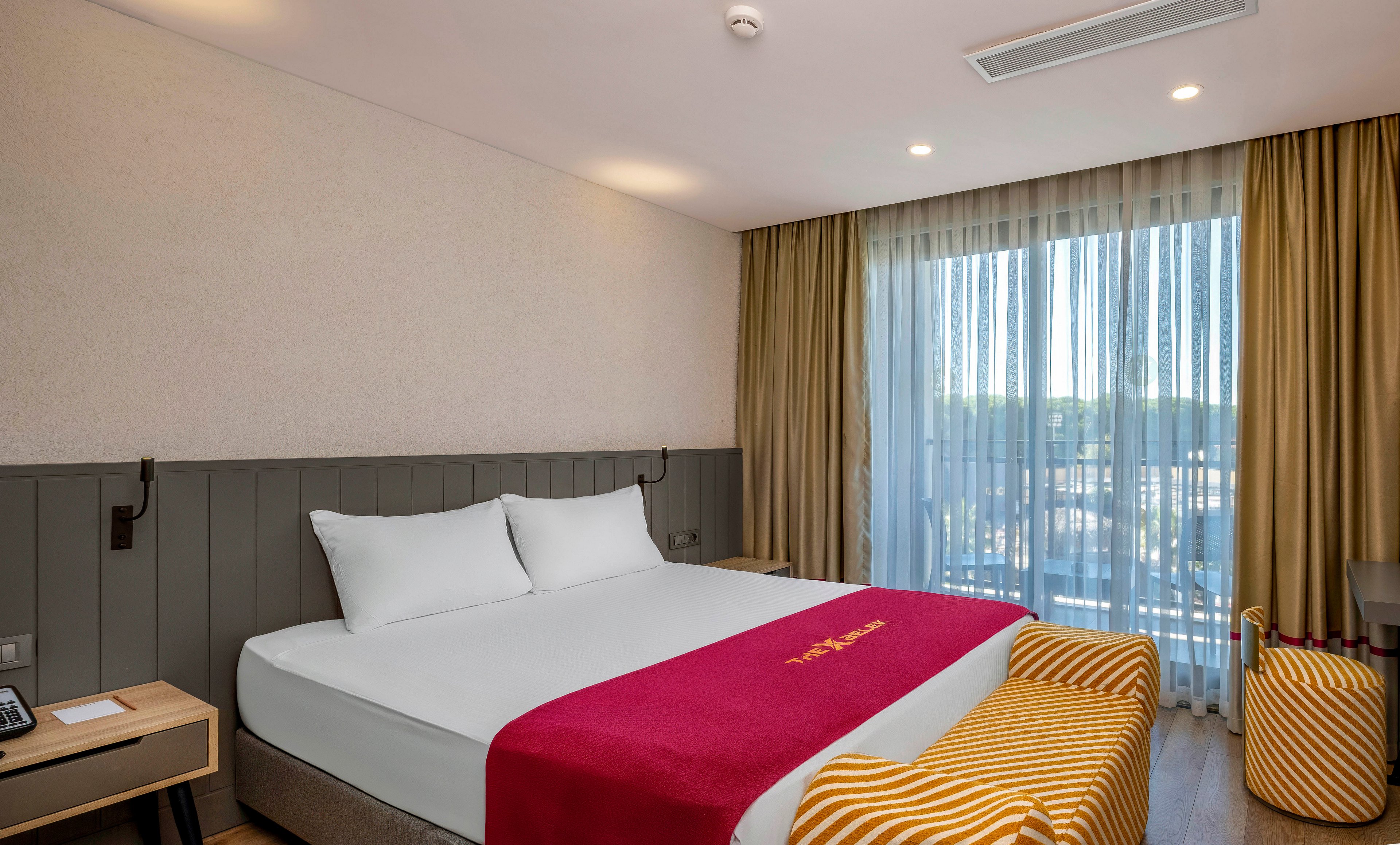 A contemporary hotel room features a large king-sized bed with crisp white linens and a deep red runner displaying gold text. The space is bright, with natural light streaming through floor-to-ceiling sheer curtains. The room includes a striped yellow and white bench and chair, wood flooring, and neutral walls, creating a welcoming and comfortable atmosphere. The bedside table holds a phone and notepad, and the overall mood is clean and inviting.