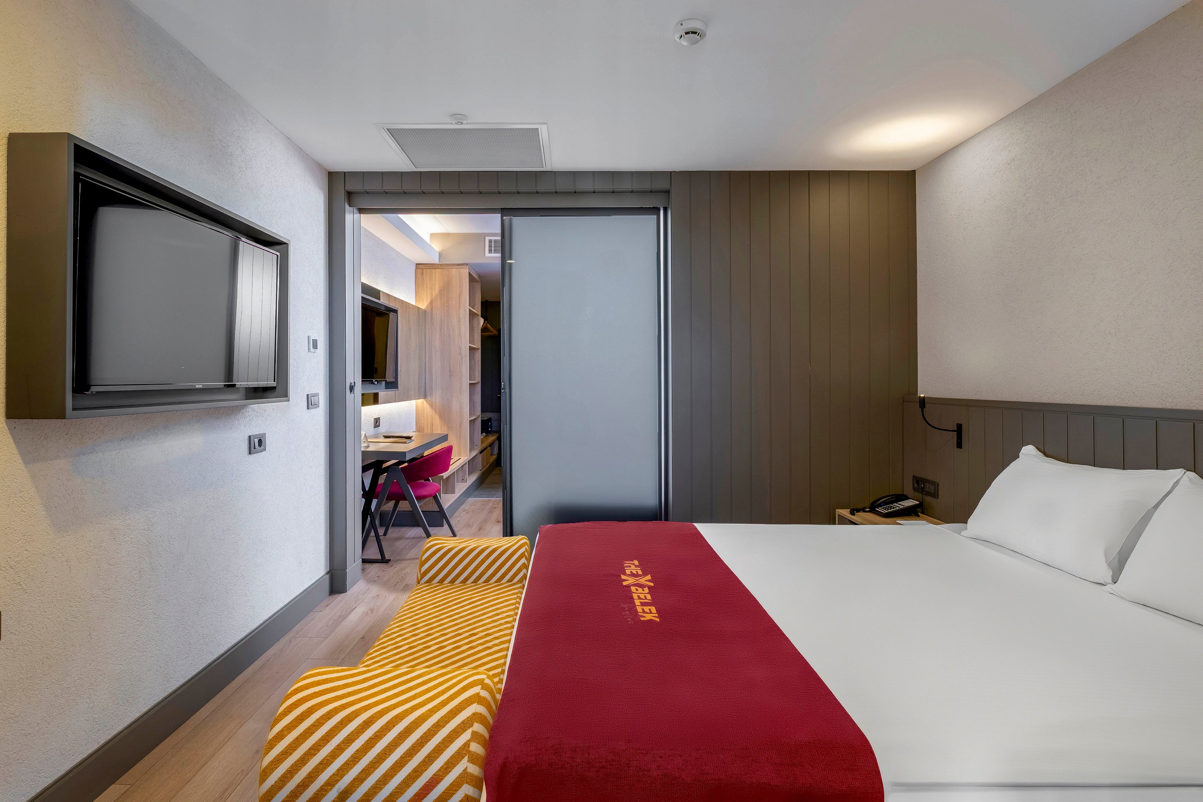 A contemporary hotel room features a neatly made bed with a red runner displaying gold text, a wall-mounted flat screen television, and a workspace with a chair visible through a sliding glass partition. The room is decorated in neutral tones with pops of color from the striped bench and red accents. The setting is clean and well-lit, creating a welcoming and comfortable atmosphere.
