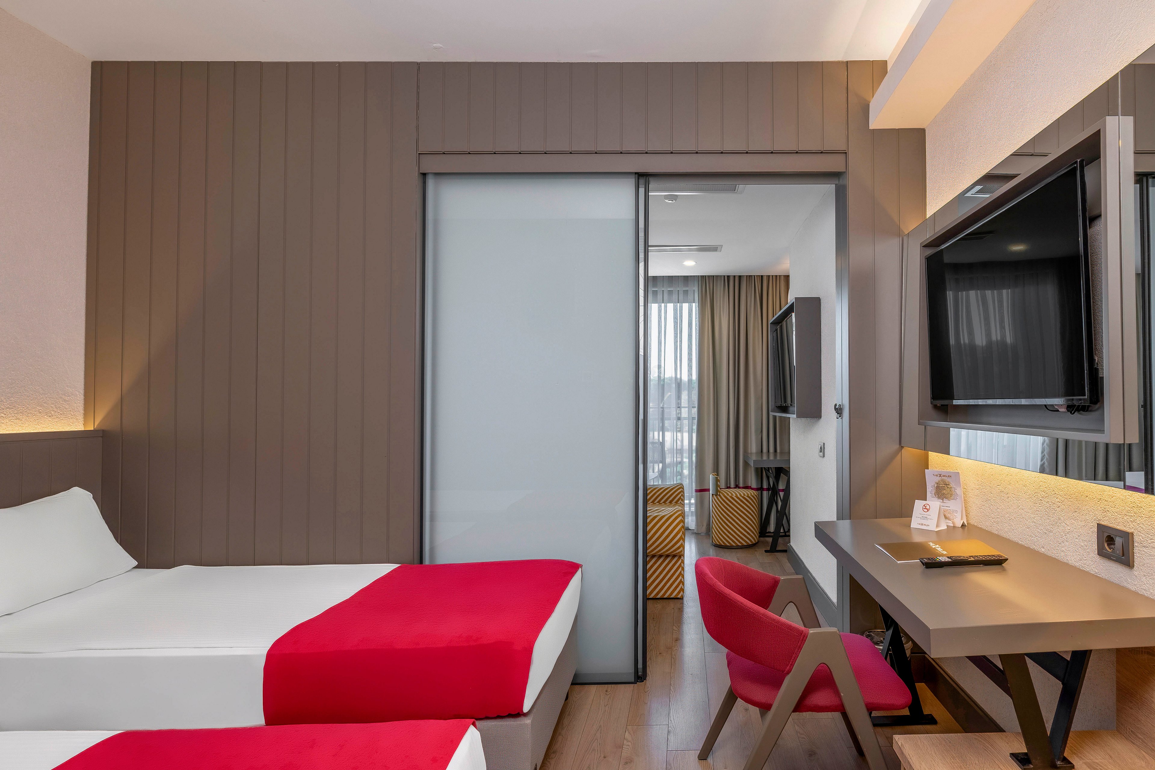 A contemporary hotel room features two twin beds with crisp white linens and bold red accents. The space includes a sleek desk area with a red chair, a wall-mounted television, and a sliding frosted glass door leading to a sitting area. Natural light streams in through large windows, highlighting the neutral palette and clean lines of the interior.
