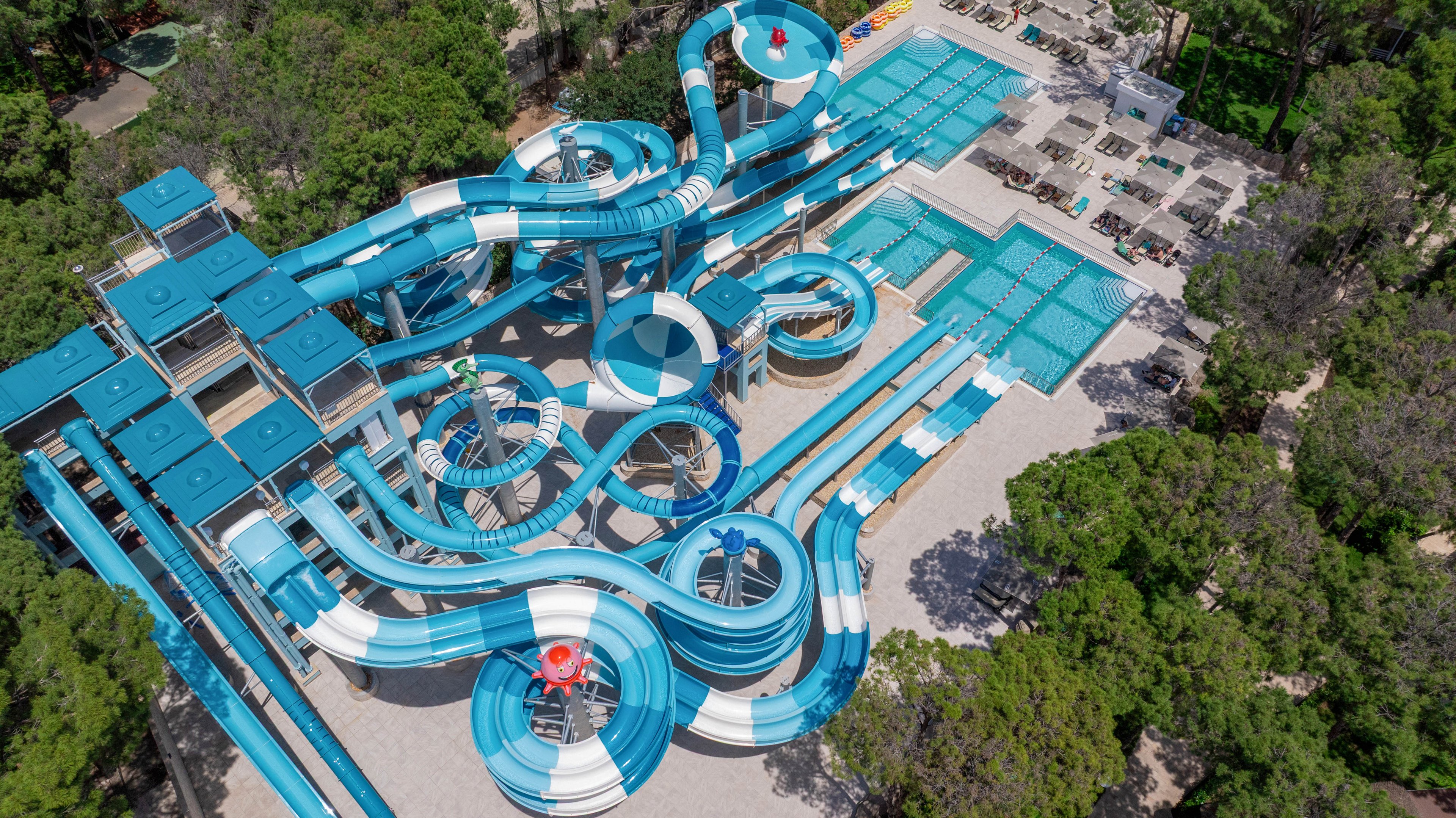 A complex network of blue and white water slides winds through an outdoor water park surrounded by trees. Multiple twisting tubes, steep slides, and circular structures connect to several rectangular swimming pools. Elevated platforms and stairways provide access to the slides, while sun loungers and seating areas are arranged around the pools. The scene is bright and colorful, emphasizing recreation and summer fun in a forested setting.