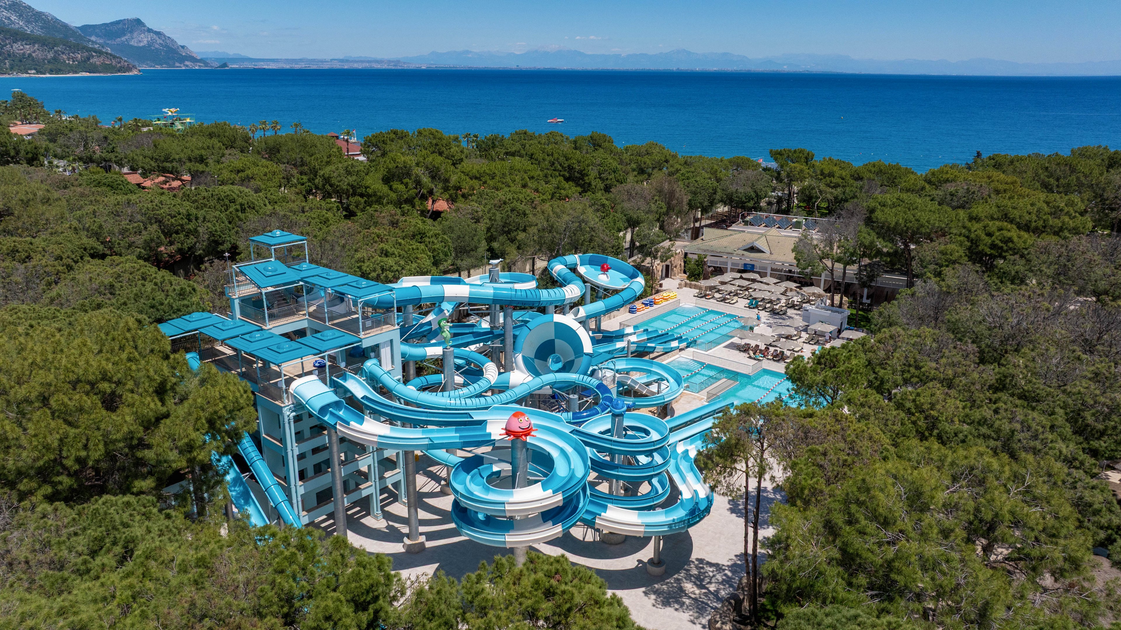 A large blue water slide complex is nestled among dense green trees near a coastline. Multiple intertwining slides and pools form the centerpiece of the water park, with observation platforms rising above the structure. The park is situated beside a bright blue sea, with distant mountains visible along the horizon. Clear skies and vibrant colors suggest a sunny, warm vacation destination.