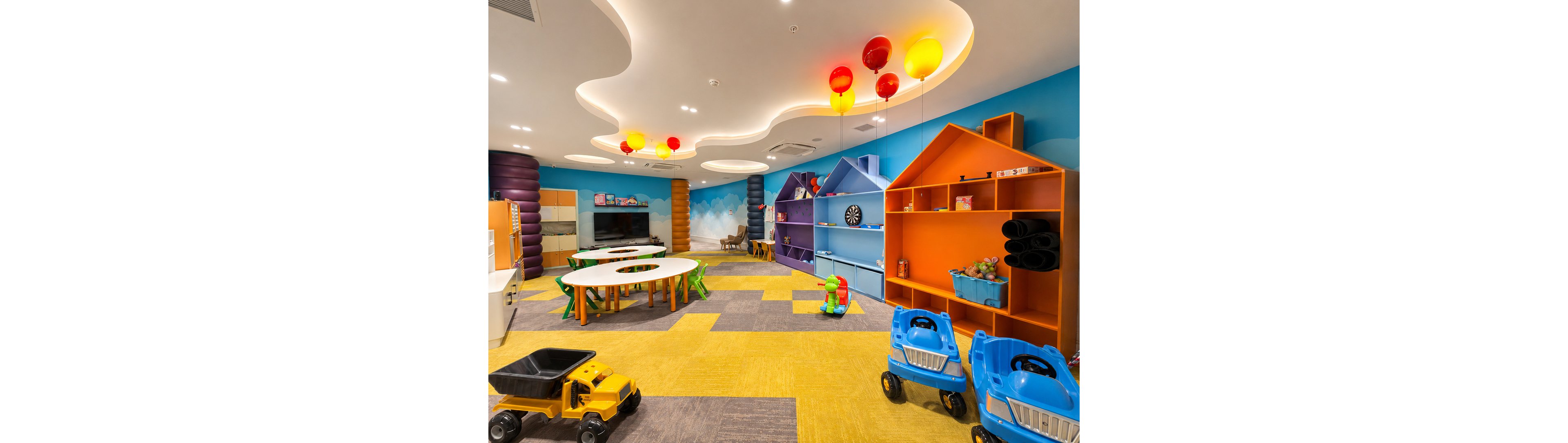 A bright, colorful indoor playroom is filled with toy vehicles and child-sized furniture. The space features house-shaped shelving units, a round activity table, and a variety of toys neatly arranged. Playful ceiling lights and balloon-like fixtures hang overhead, while a whimsical wall mural adds to the fun atmosphere. The room appears clean, organized, and designed for young children’s activities and play.
