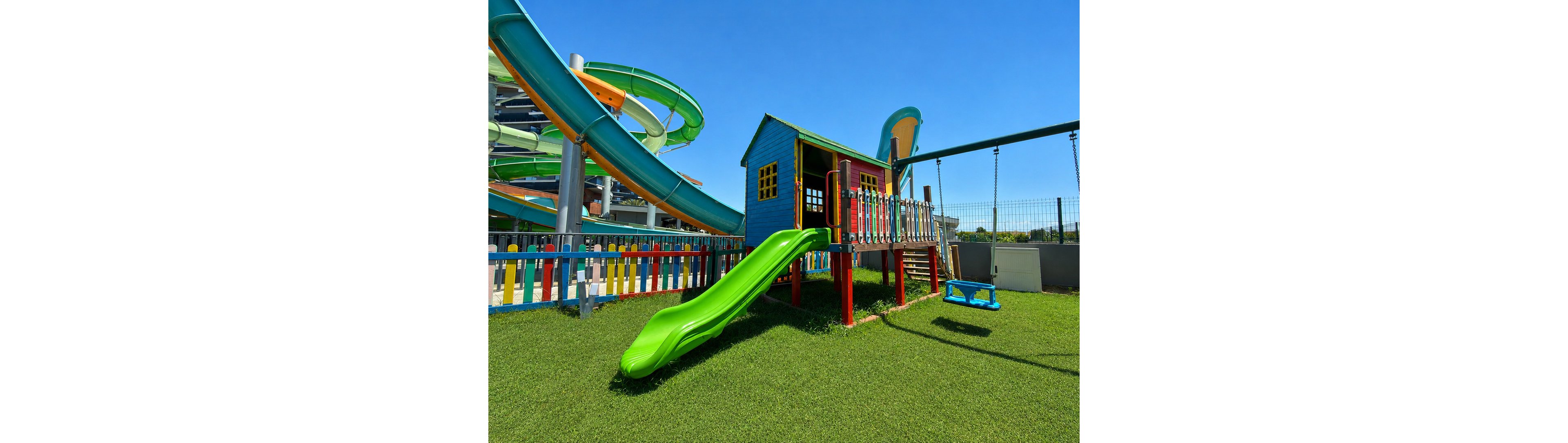 A bright playground structure with a green slide and small wooden playhouse stands on a neatly trimmed grass surface. Next to it, a blue swing hangs from a metal frame. In the background, large twisting water slides and a multicolored fence add to the fun, recreational atmosphere. The scene is set under a clear blue sky, suggesting a sunny day at an outdoor park or water park.