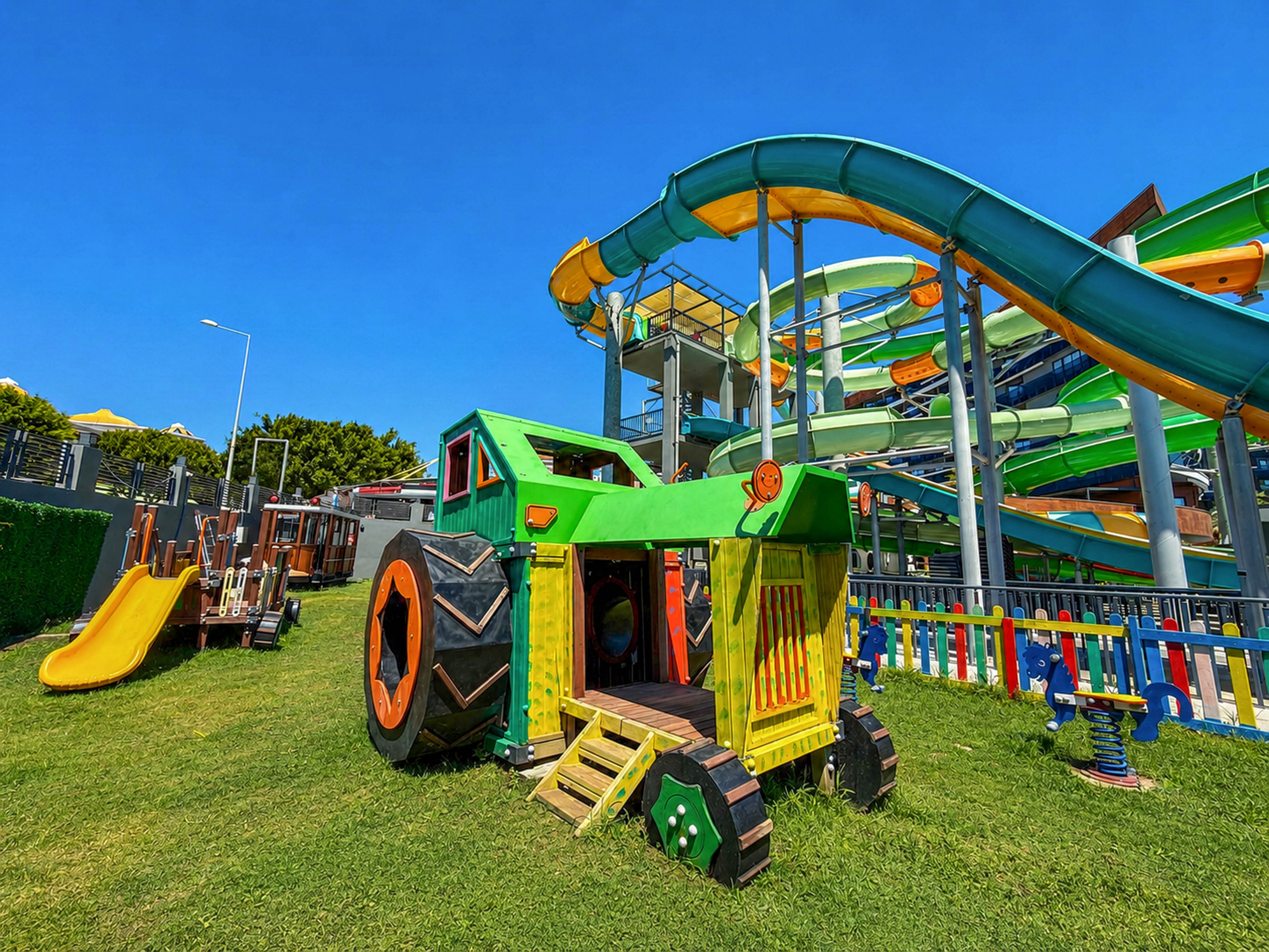 A bright green toy tractor structure stands on a grassy playground under a clear blue sky. Nearby, a yellow slide and other wooden play equipment provide additional activities for children. In the background, large twisting water slides with yellow and green sections rise above the scene. A colorful fence separates the playground from the water park area, creating a lively and family-friendly environment.