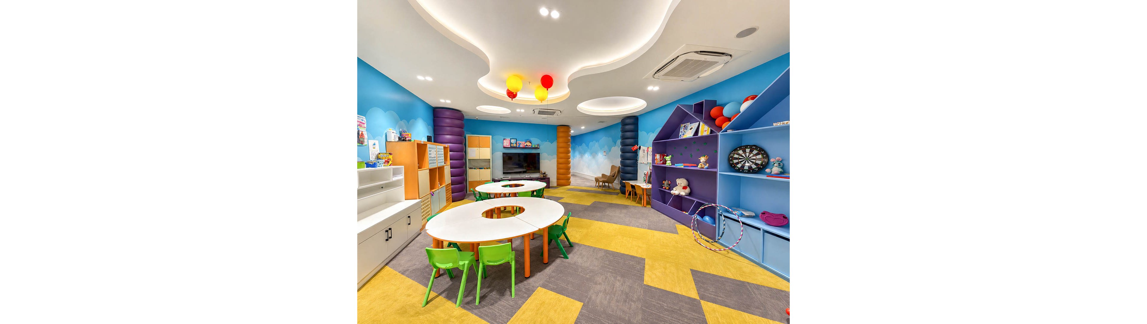 Brightly colored children's playroom featuring round white tables with small green chairs in the center. The room has playful wall designs with clouds, balloons, and bold geometric shelving filled with toys and books. A dartboard, hula hoops, and stuffed animals add to the fun, activity-focused atmosphere. Overhead recessed lighting and ceiling fixtures create a clean, modern look suitable for a daycare or preschool setting.