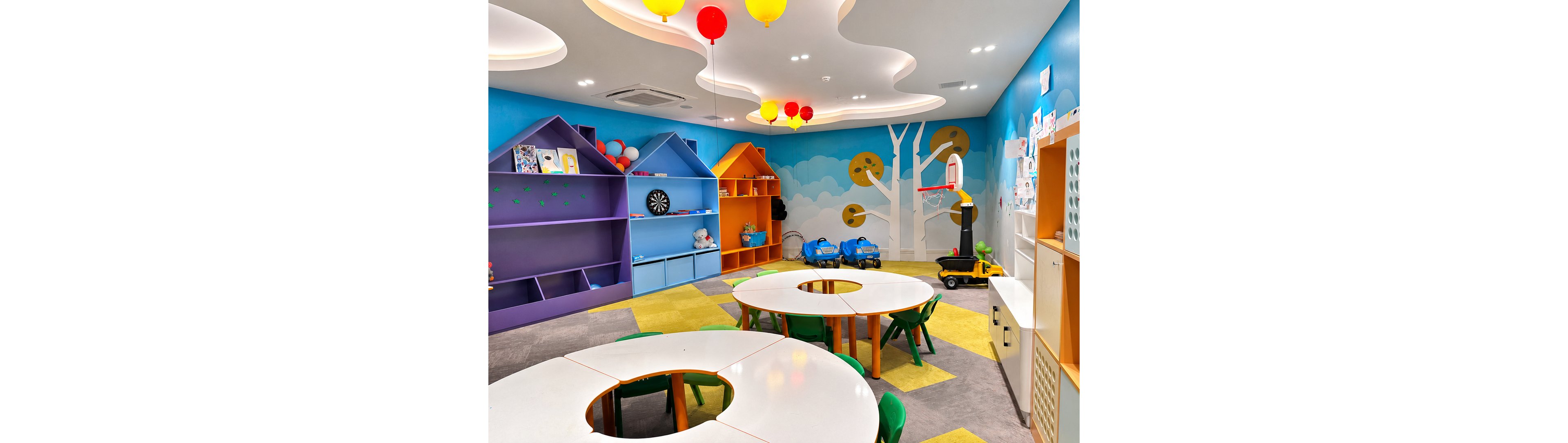 A bright preschool classroom features round white tables with small green chairs arranged for group activities. Colorful built-in shelves shaped like houses line the wall, holding books, toys, and a dartboard. The walls are painted with playful tree and cloud illustrations, and the ceiling has wavy panels with hanging red and yellow balloons. Toy cars and a small basketball hoop are placed along the side, creating a fun and inviting learning environment.