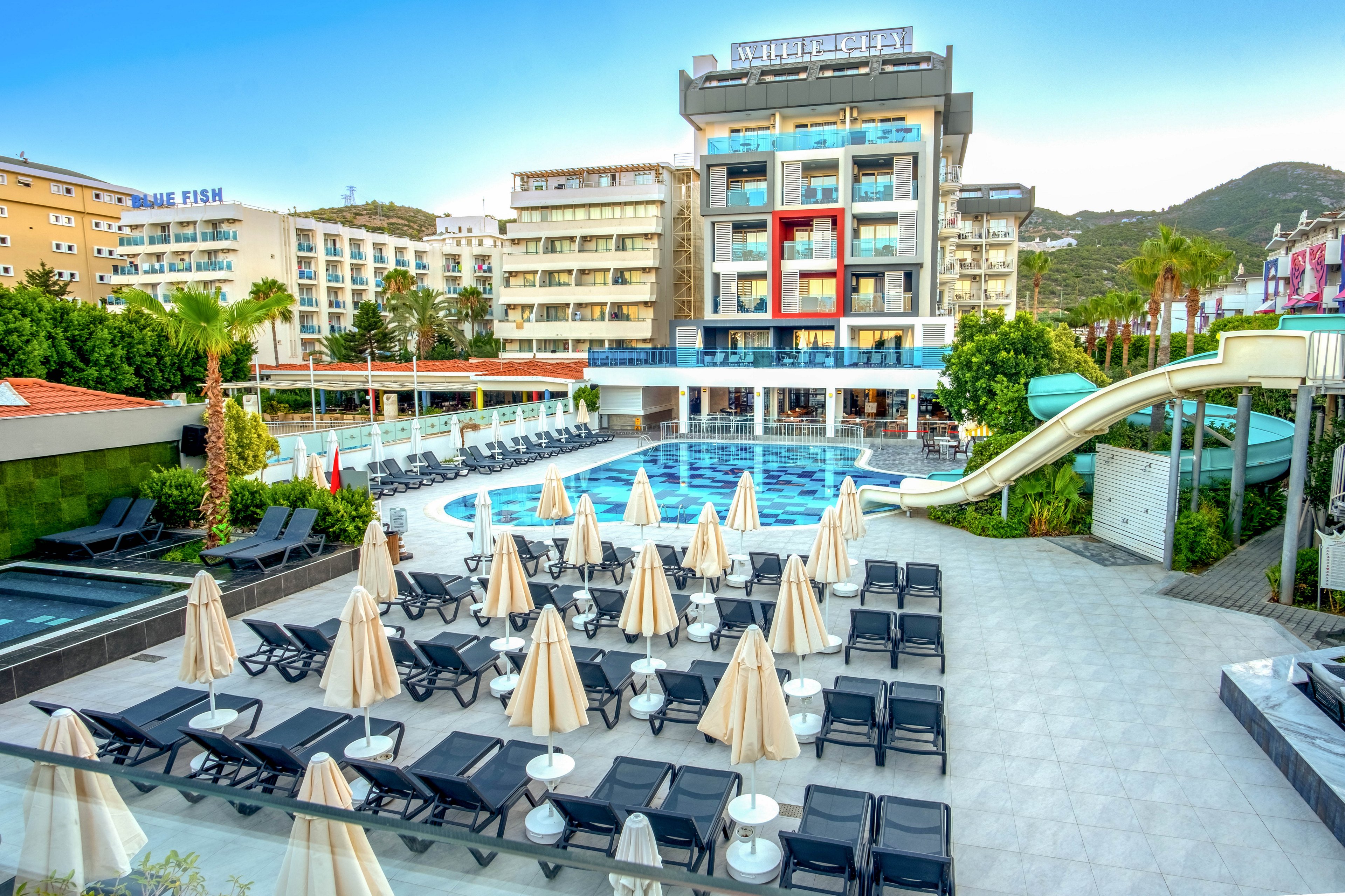 A contemporary multi-story resort hotel overlooks a large outdoor swimming pool lined with sun loungers and closed umbrellas. The scene is set in a sunny urban coastal area with surrounding buildings and distant green hills. A curved waterslide leads into the pool, adding a playful element to the otherwise calm and orderly terrace. The bright blue water, clean white tiles, and glass balconies create a fresh, inviting vacation atmosphere.