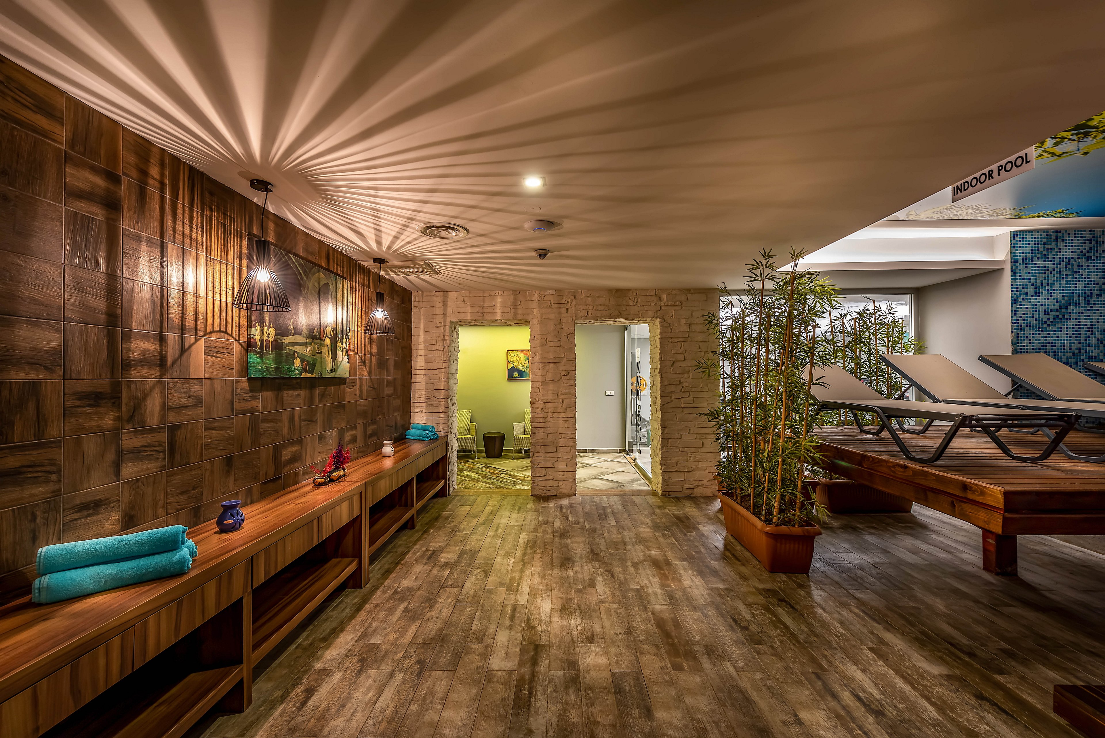 A contemporary spa lounge features wooden floors, warm lighting, and a long console table with neatly folded turquoise towels. Comfortable lounge chairs are arranged on a raised wooden platform near potted plants, creating a tranquil relaxation zone. The walls combine textured wood and light stone finishes, leading to an open doorway and additional rooms in the background. The overall ambiance is calm and inviting, ideal for wellness, rest, and rejuvenation.
