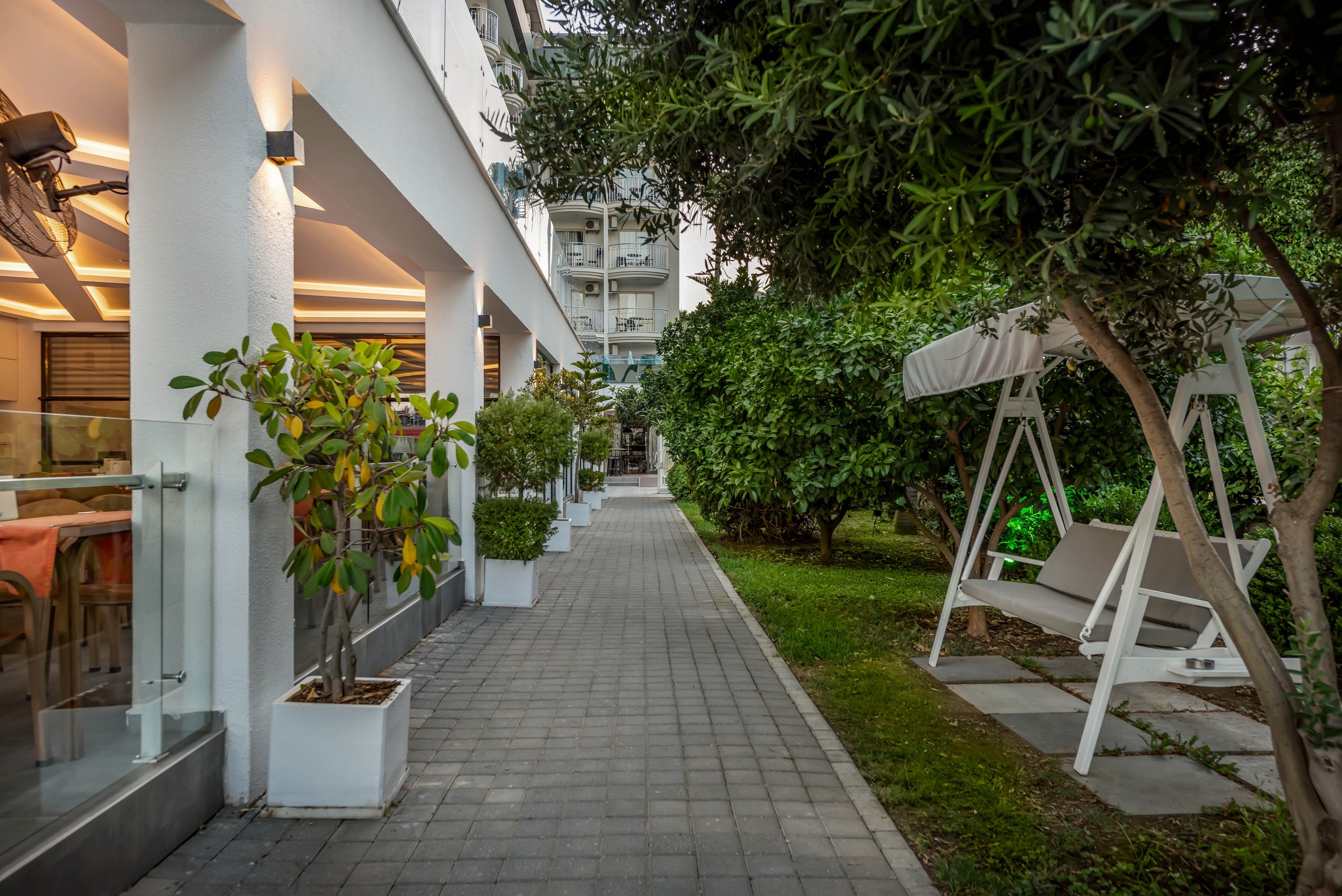 A paved walkway runs between a modern building terrace and a lush green garden. On the right side, a white swing bench with a canopy sits among trees and shrubs. The building features large windows, warm interior lighting, and potted plants along the path. The scene feels calm and inviting, suggesting an outdoor relaxation or hospitality area in an urban setting.