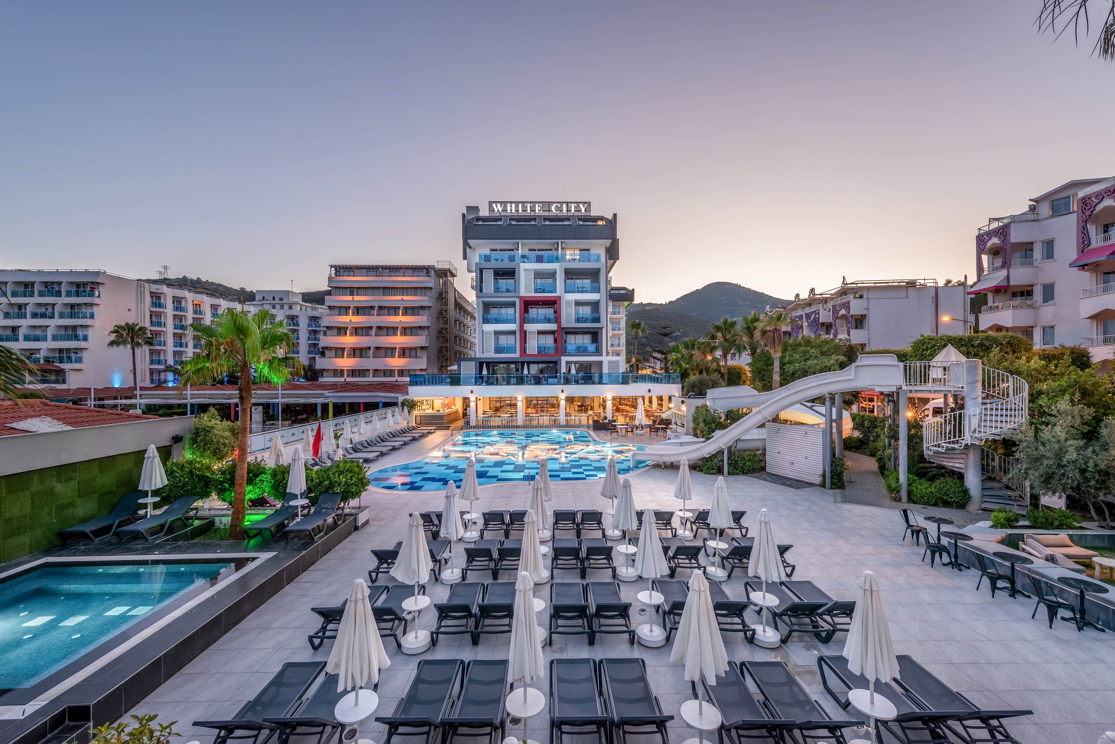 A contemporary multi-story hotel stands at the center of a coastal resort complex at sunset. In the foreground, rows of sun loungers and closed umbrellas surround a large swimming pool area. A waterslide curves into the main pool, while a smaller rectangular pool sits to the side. Surrounding buildings, palm trees, and distant hills frame the tranquil vacation setting under a clear evening sky.