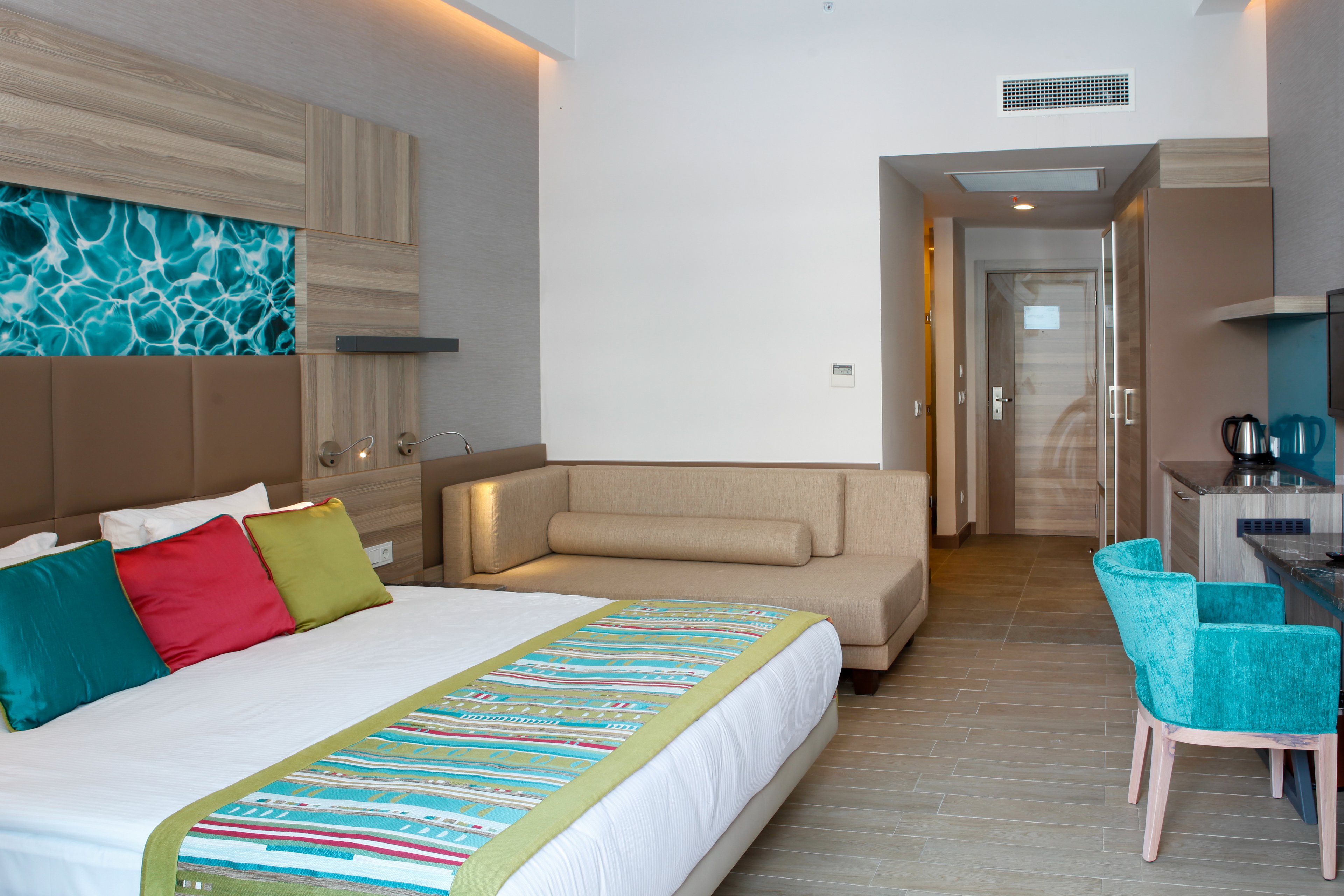 A contemporary hotel room featuring a large bed with vibrant pillows and a patterned bed runner. The setting includes a cozy seating area, a desk with teal chairs, and a kettle on the counter. The room is well-lit with natural and artificial lighting, showcasing a turquoise accent wall with a water-inspired design.