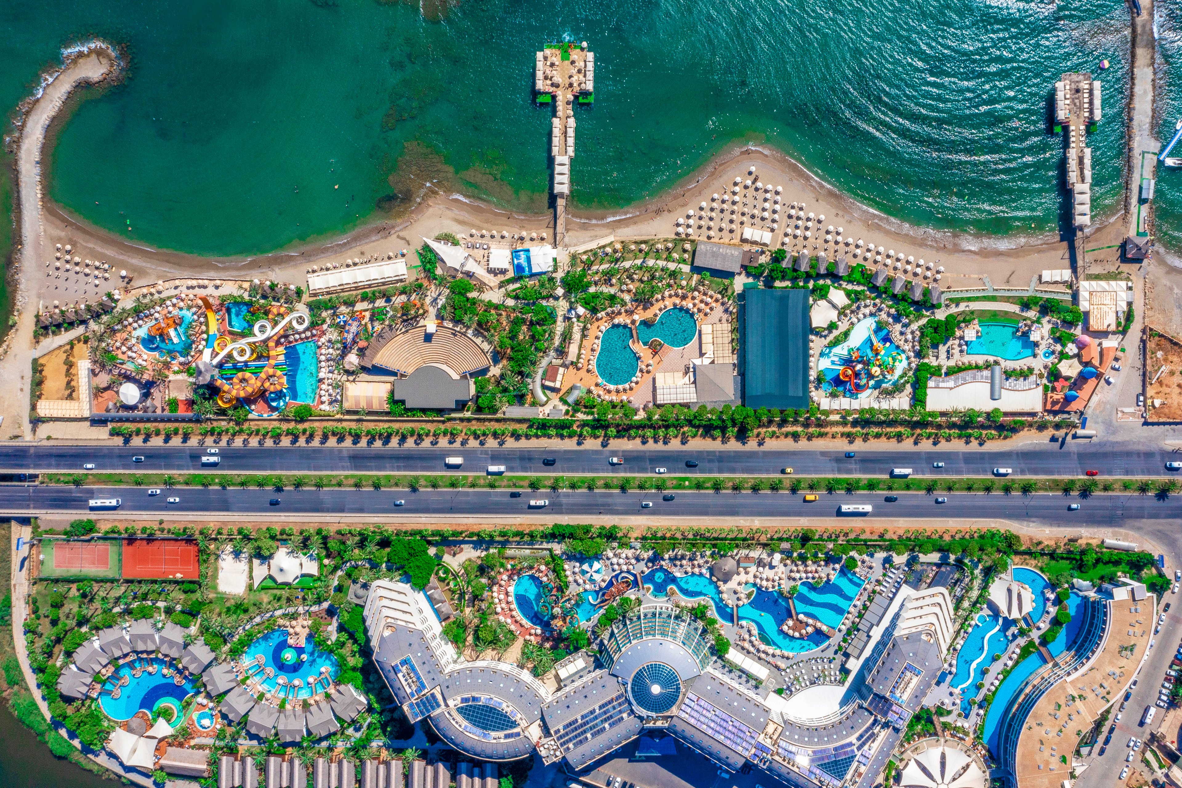 A vibrant seaside resort complex is captured from above, showing multiple swimming pools, water slides, and landscaped areas. A main road runs horizontally through the center of the image, separating the hotel buildings from the beachfront zone. On the beach side, rows of sun loungers and umbrellas line the sandy shore next to calm green-blue sea water. The scene is colorful and busy, emphasizing leisure, vacation, and waterfront recreation.