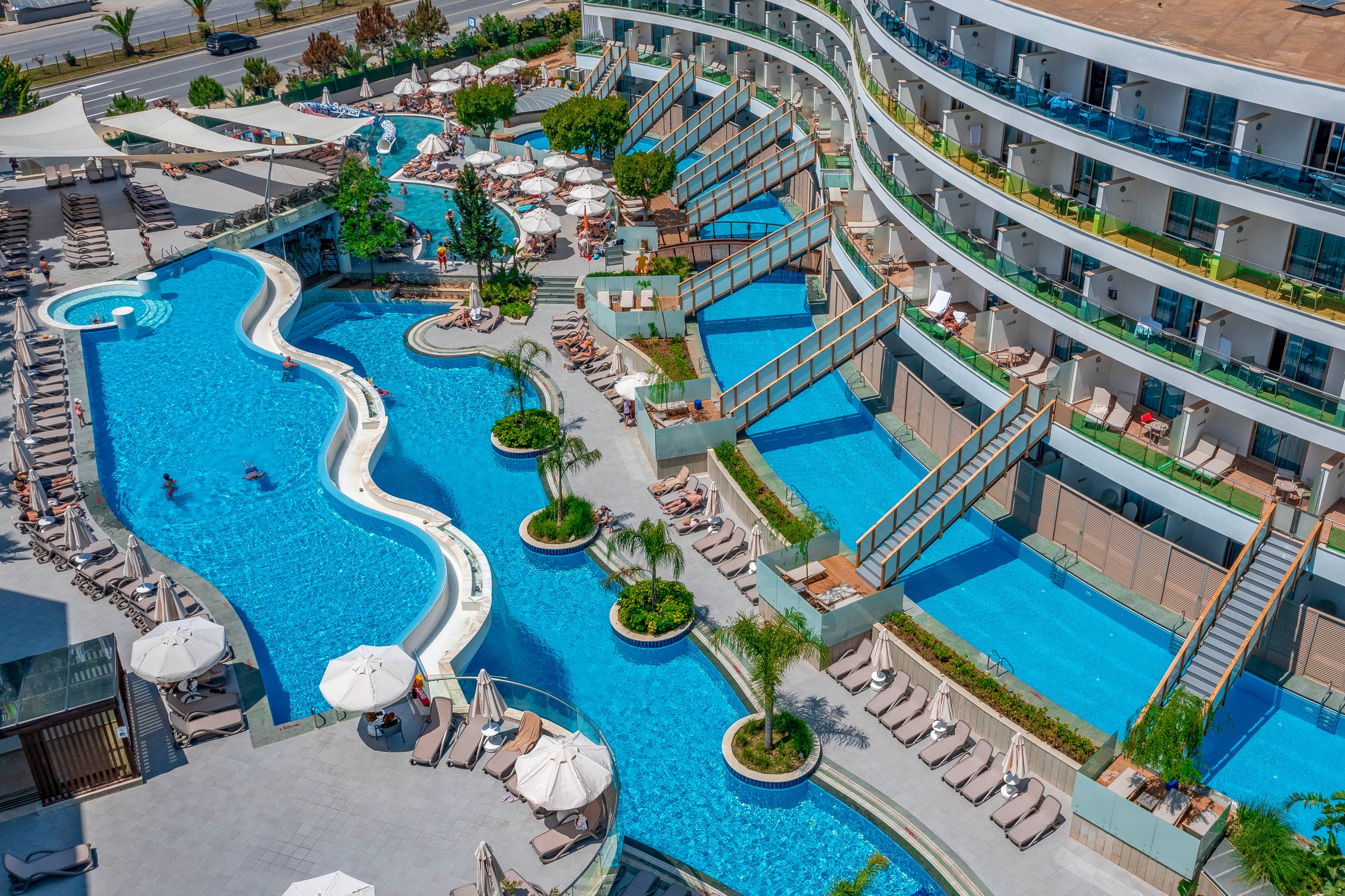 A contemporary resort features a large, winding outdoor swimming pool surrounded by sun loungers and umbrellas. Private terrace pools cascade down from the curved hotel building, each separated by glass railings and steps. Guests relax in the main pool and on deck chairs under clear, sunny skies. Palm trees, landscaped greenery, and shaded seating areas create a luxurious vacation atmosphere near a coastal roadway.