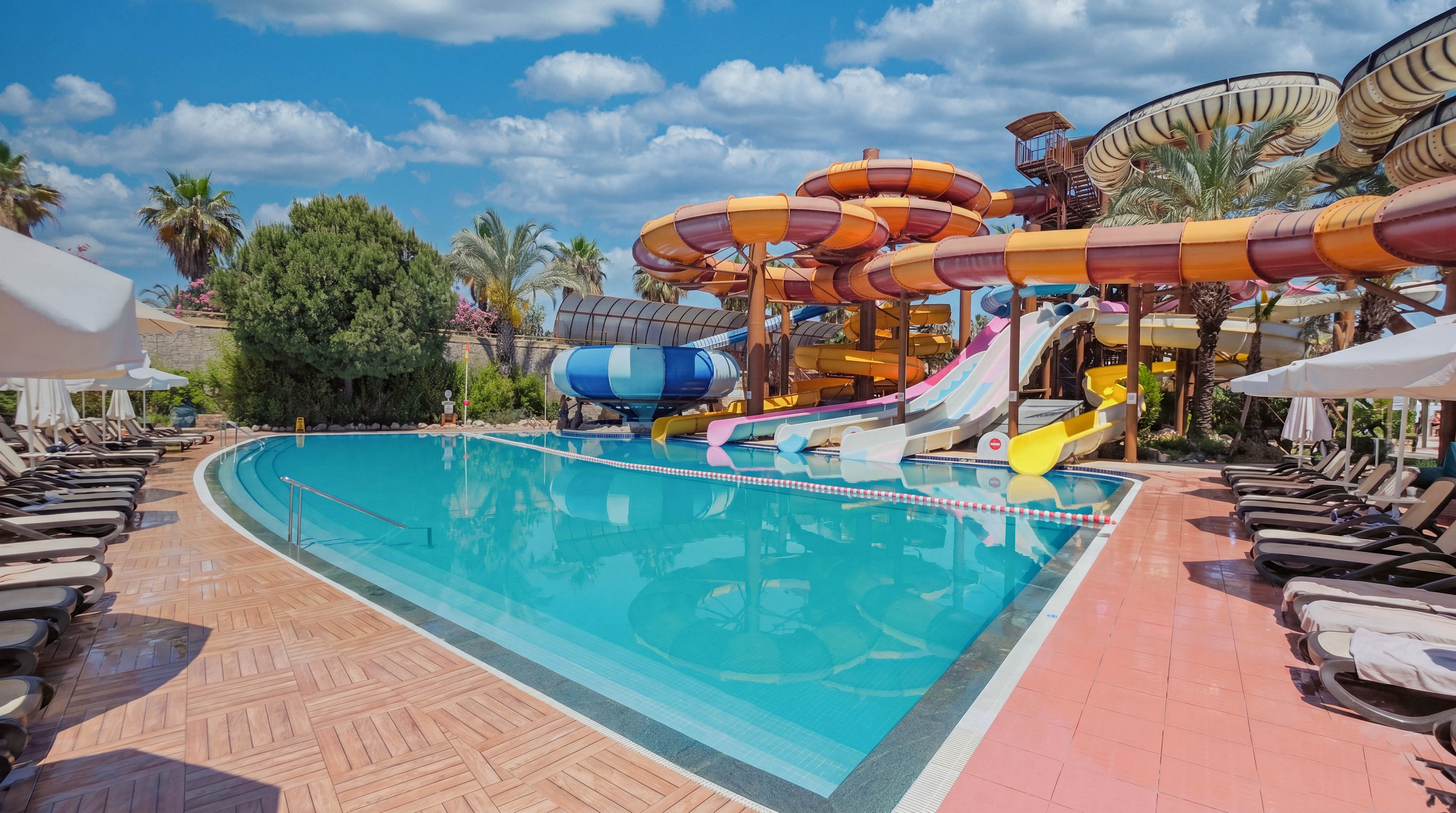 A large outdoor swimming pool sits in front of a complex of colorful water slides at a resort. Empty lounge chairs line the tiled pool deck on both sides, suggesting a quiet moment before guests arrive. Palm trees and lush greenery surround the area, adding a tropical atmosphere under a bright blue sky with scattered clouds. The scene conveys a fun, family-friendly vacation destination focused on water activities and relaxation.