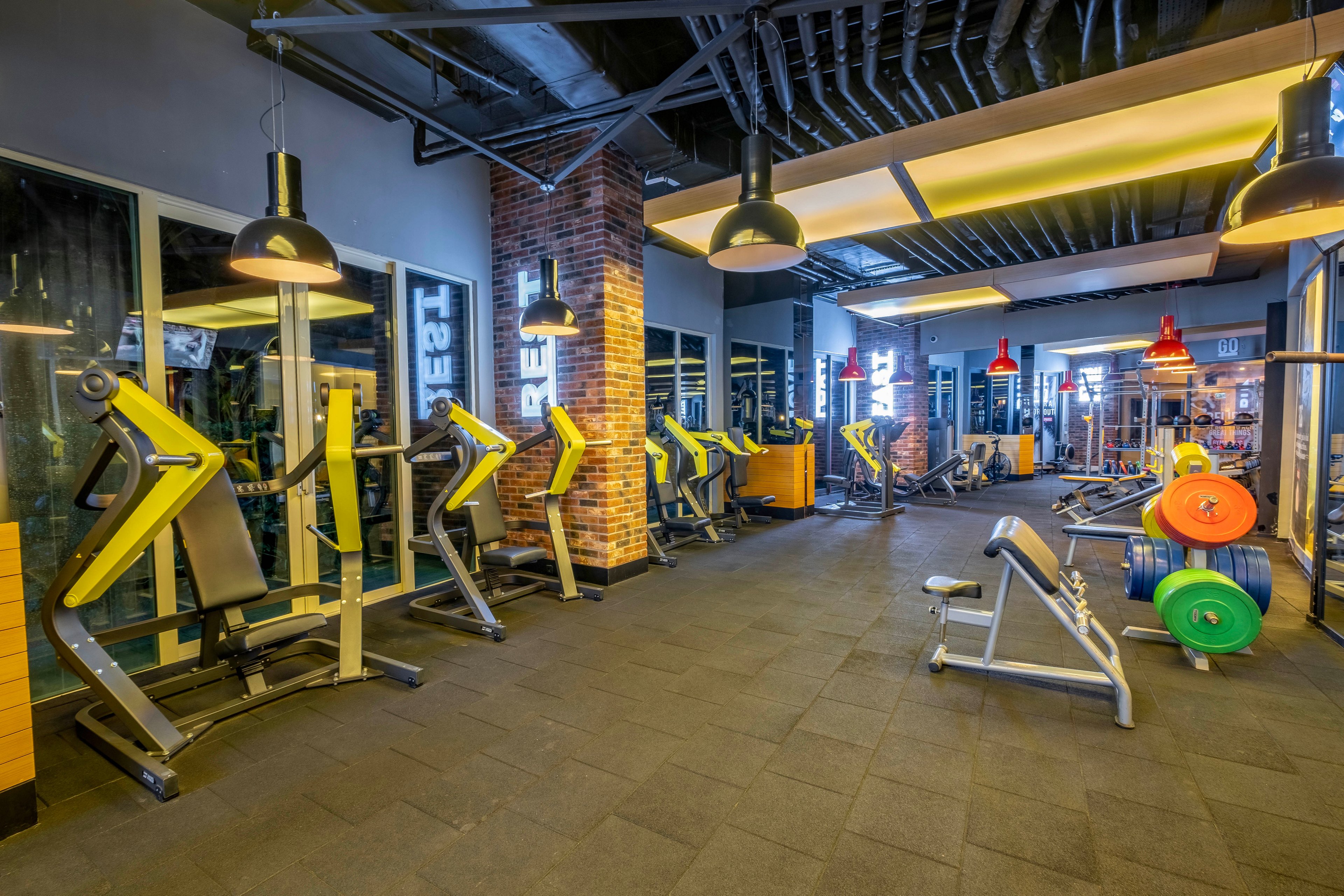 A brightly lit modern gym interior features multiple strength training machines lined up along large windows. Colorful weight plates are stacked on racks and barbells in the foreground, with benches and additional equipment visible further back. Industrial-style lighting, exposed ceilings, and brick columns create a contemporary fitness studio atmosphere. The space appears clean, organized, and ready for workouts, but no people are present.