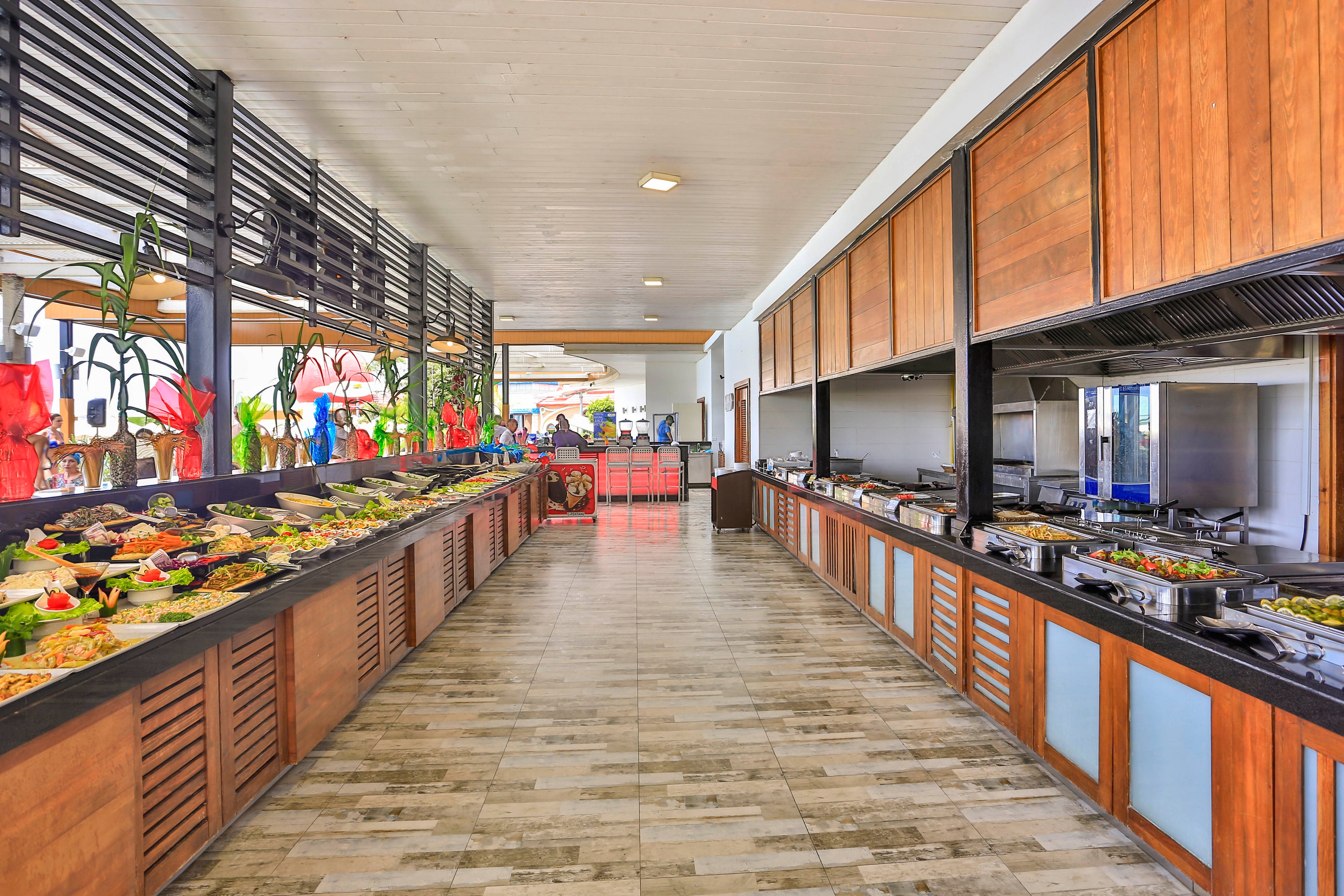 A long self-service buffet counter is filled with a wide variety of dishes and salads. The setting appears to be a bright, modern hotel or resort dining area with wooden cabinetry and tiled flooring. Food trays and platters are neatly arranged along both sides of the corridor-like space. Decorative plants and colorful wrapped items add visual interest near the serving area. No people are present, emphasizing the layout and abundance of the buffet spread.