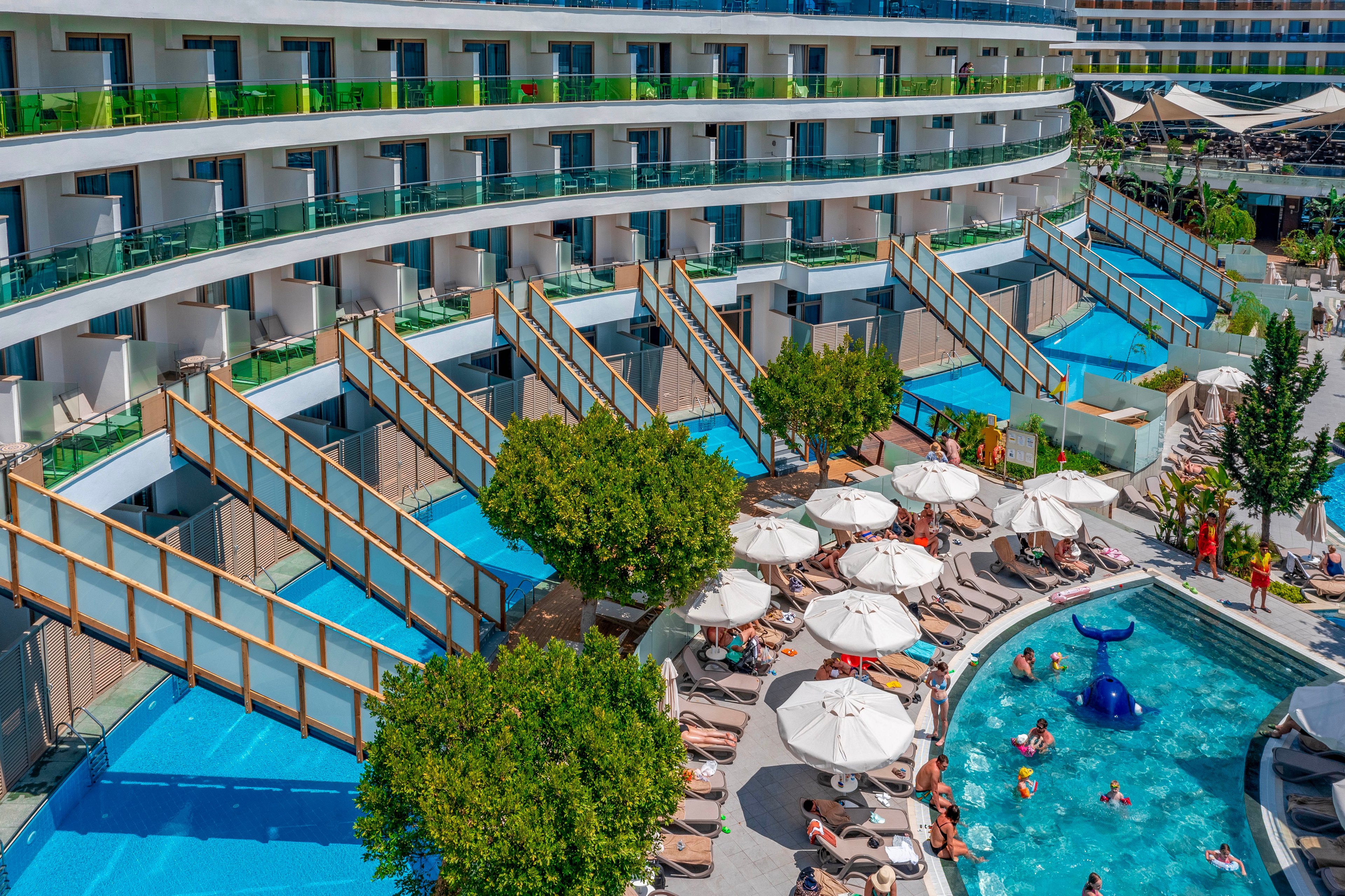 A contemporary resort hotel features multiple floors of rooms opening onto cascading private pool terraces. In the foreground, guests relax around a large outdoor swimming pool lined with white sun loungers and umbrellas. Families and children play in the water near a blue whale-shaped slide and other pool toys. Green trees, bright railings, and clear blue water create a vibrant, sunny vacation atmosphere.