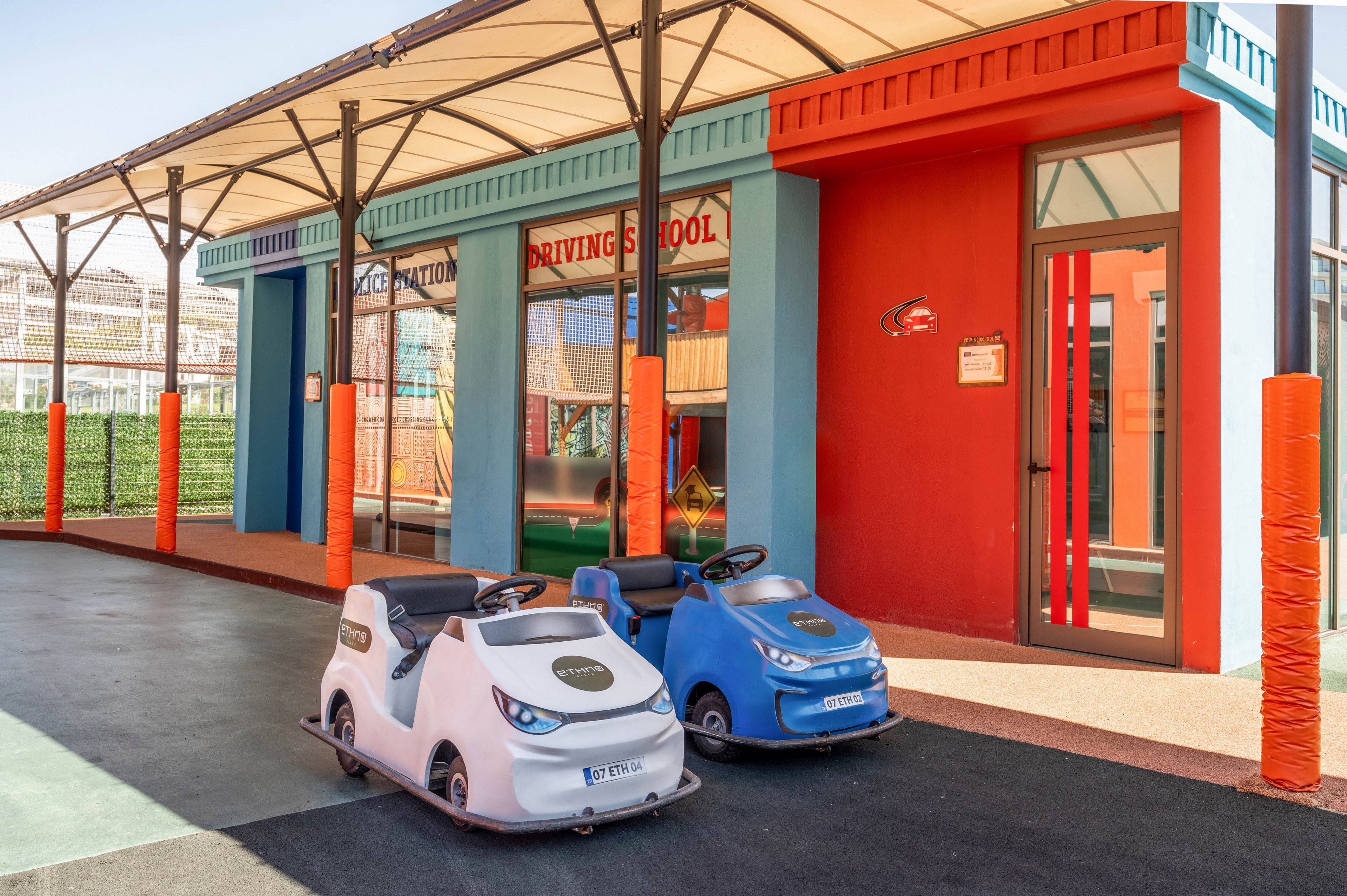 Two small electric toy cars are parked in front of a brightly colored building at a children's driving school. The white and blue cars sit on a smooth outdoor track under a covered walkway. The building features bold red, blue, and orange walls with large windows and doors. Sunlight enhances the playful atmosphere of this educational play area.
