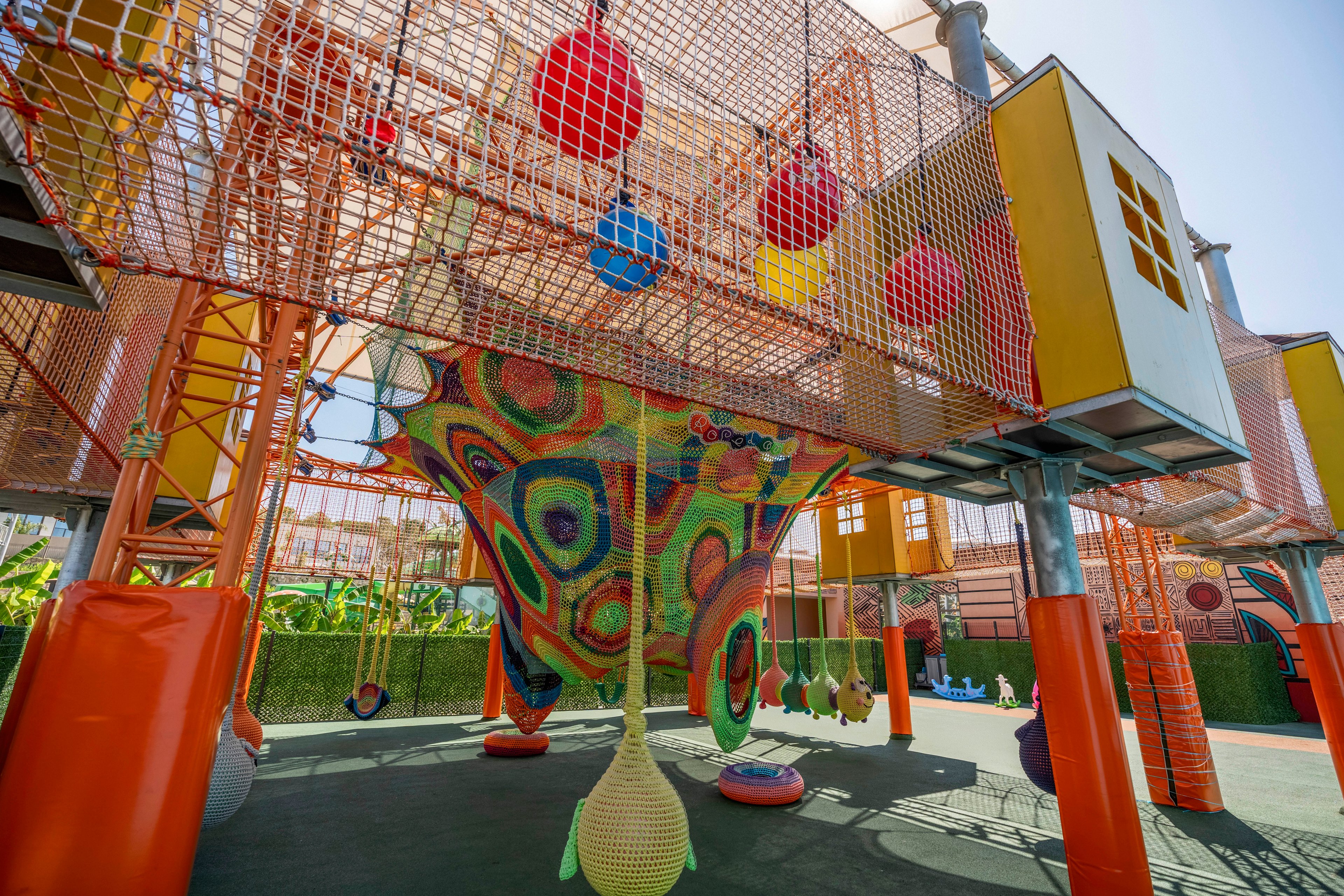 A vibrant outdoor playground features an elaborate climbing structure with nets, ropes, and hanging pods. Bright orange frames support elevated walkways and enclosed play towers. Multicolored woven nets and suspended balls create a dynamic, playful environment. Sunlight and surrounding greenery emphasize the open-air, family-friendly setting.