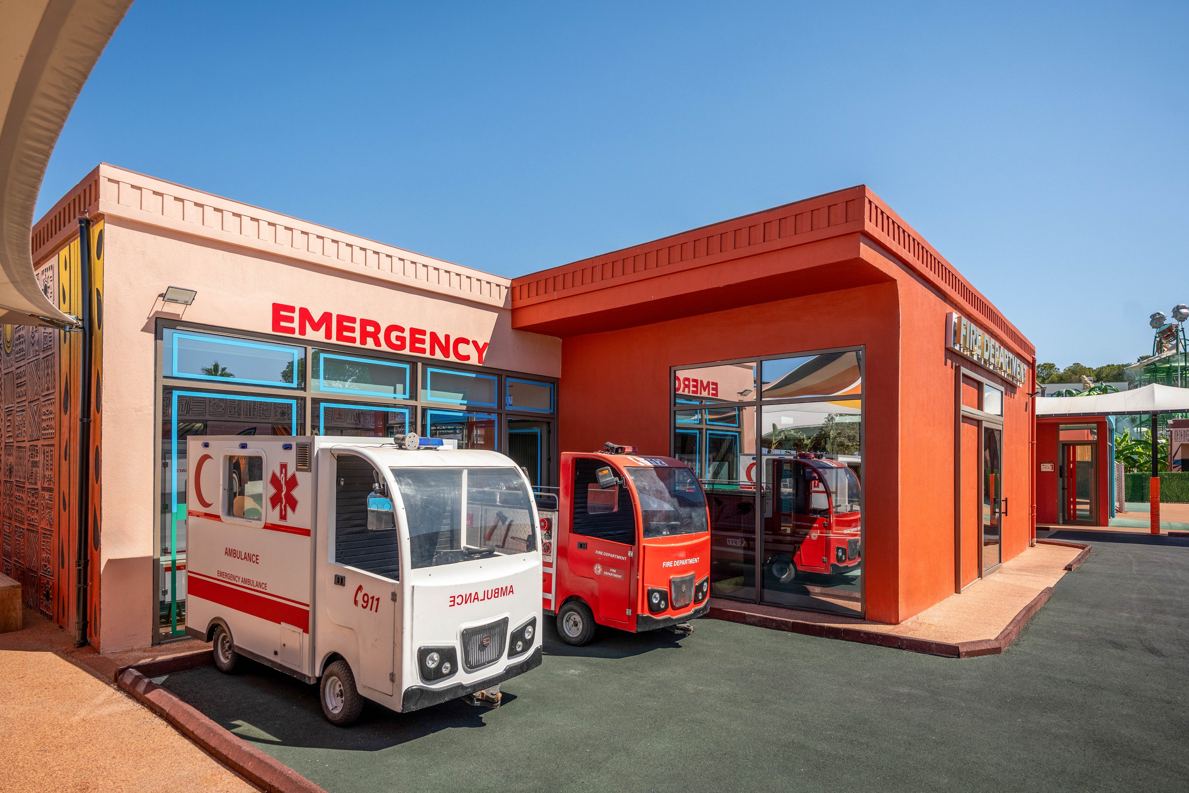 Small toy ambulances are parked outside a brightly colored emergency play clinic. The building features bold red and orange walls with large windows reflecting the vehicles. Clear signage reading "EMERGENCY" and "AMBULANCE" emphasizes the medical theme. The sunny outdoor setting and playful proportions suggest a children’s play area or educational space.