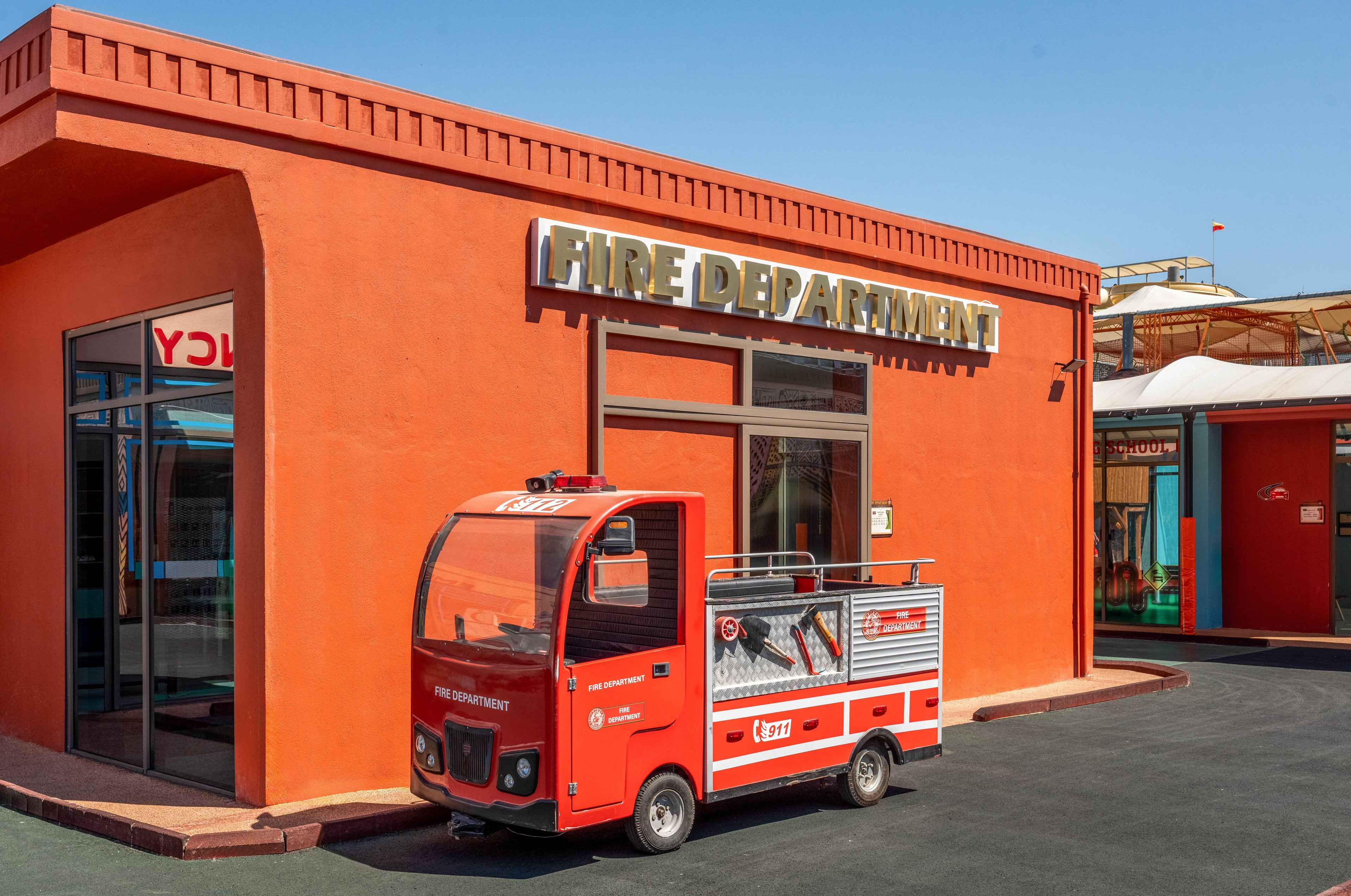 A small red fire truck is parked in front of an orange fire department building. The structure features large windows and a bold sign reading "FIRE DEPARTMENT" above the entrance. The scene appears to be part of a themed or educational area with other colorful buildings nearby. Clear blue sky and bright sunlight create a vivid, playful atmosphere.