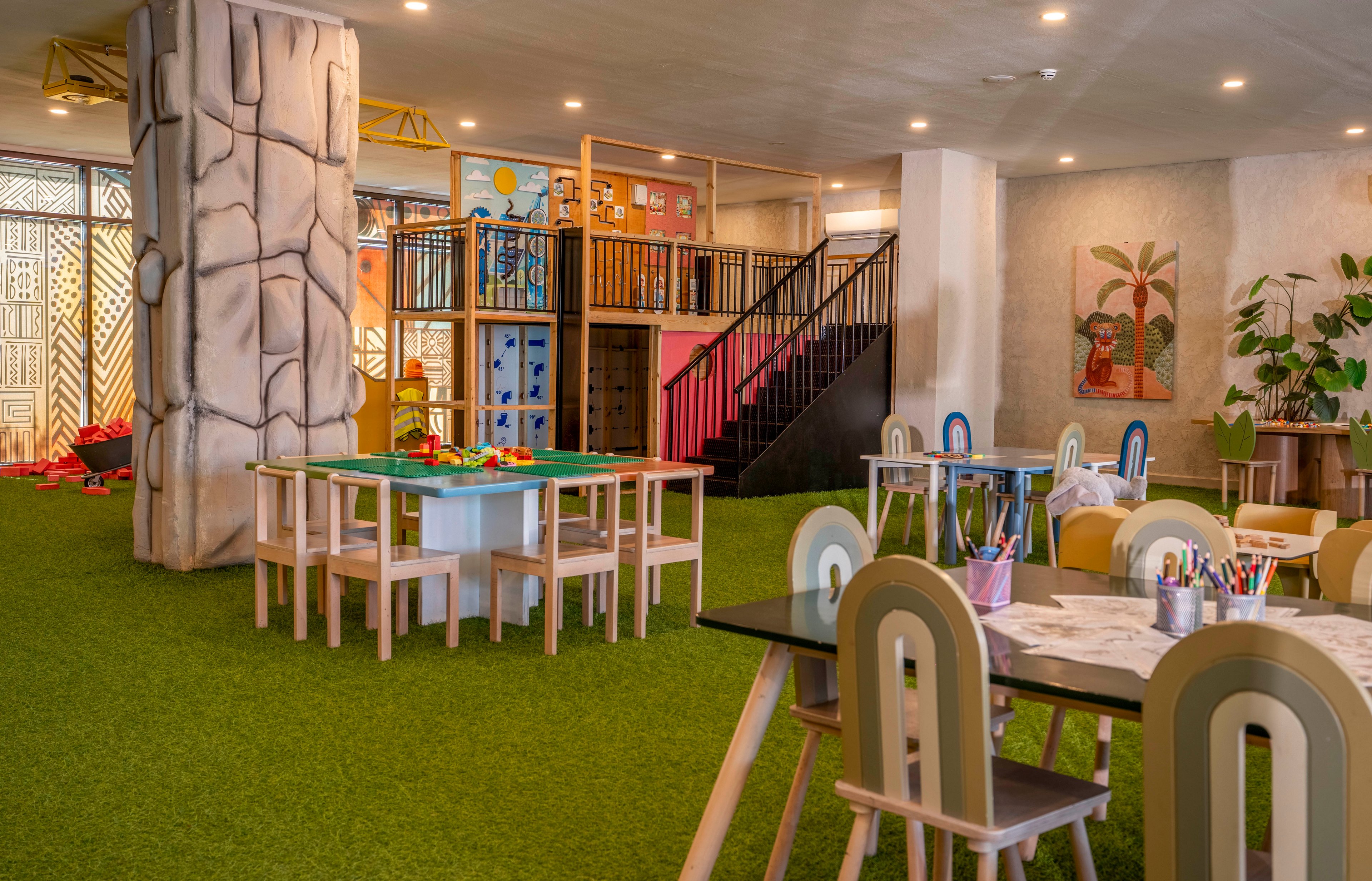 Spacious indoor children's playroom featuring small wooden tables and chairs arranged on bright green artificial grass flooring. A multi-level play structure with stairs, railings, and climbing elements stands against the back wall. Art supplies and toys are neatly placed on the tables, suggesting creative activities and playtime. Warm lighting, playful wall art, and potted plants create a cheerful and inviting atmosphere.