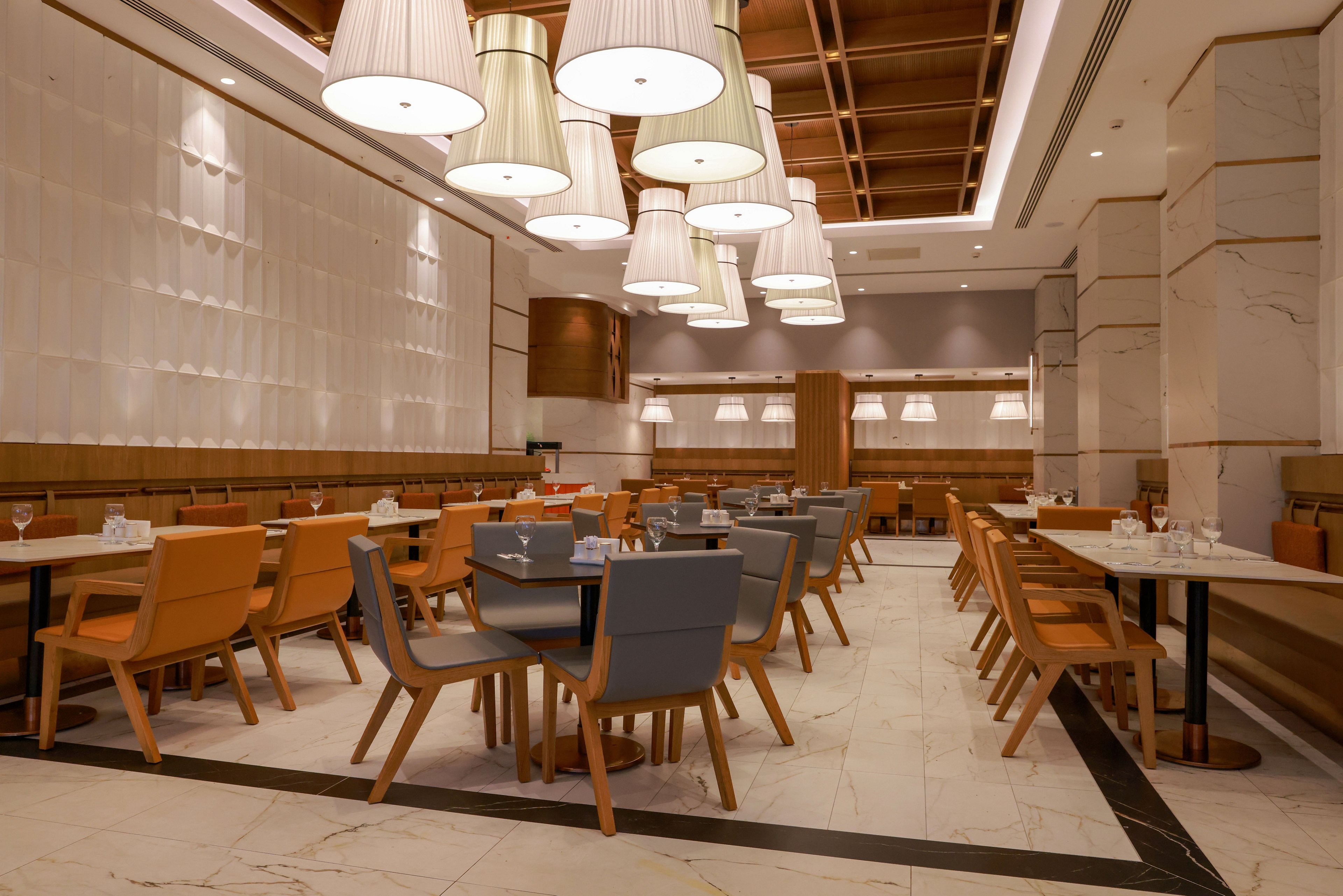 A contemporary restaurant dining area features rows of tables and chairs arranged neatly in an empty space. Warm pendant lights hang from a wooden ceiling, casting a soft glow over the marble floor and neutral-toned walls. Orange and gray upholstered chairs add color contrast to the otherwise beige and white interior. Glassware and place settings on the tables suggest the space is prepared for guests.