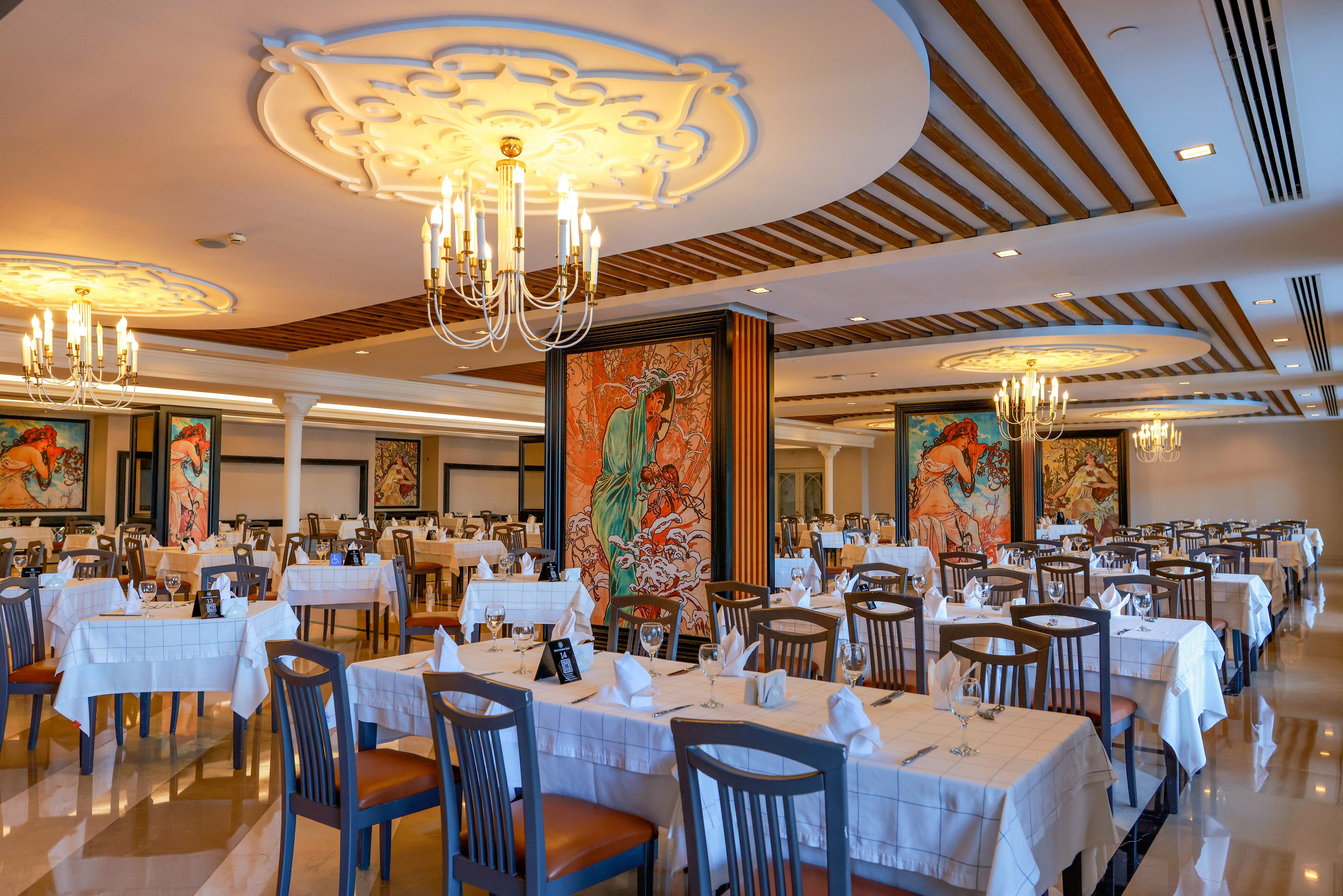 Spacious dining room with neatly arranged tables and chairs is set for guests in an upscale restaurant. White tablecloths, folded napkins, and table settings create a formal atmosphere. Large colorful paintings on pillars add artistic flair to the interior. Warm chandelier lighting and decorative ceiling details enhance the sophisticated, inviting ambiance.