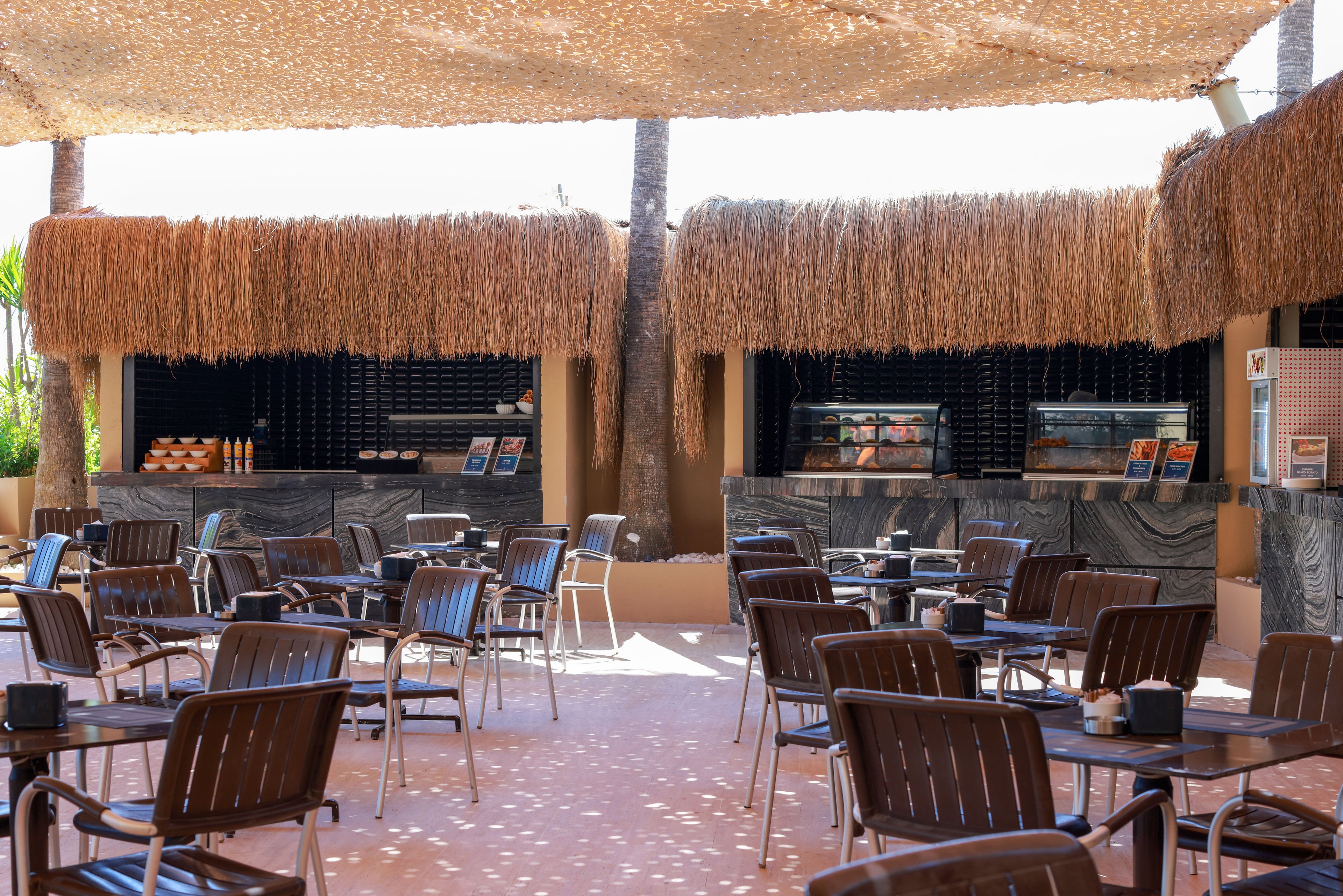 An open-air café area features multiple service counters with straw-covered roofs and shaded seating. Numerous metal and wooden chairs surround small tables arranged on a sunlit terrace. The counters display food items and menu boards behind glass, suggesting a casual dining or snack bar setting. A woven canopy overhead casts dappled light across the floor, creating a relaxed vacation atmosphere.