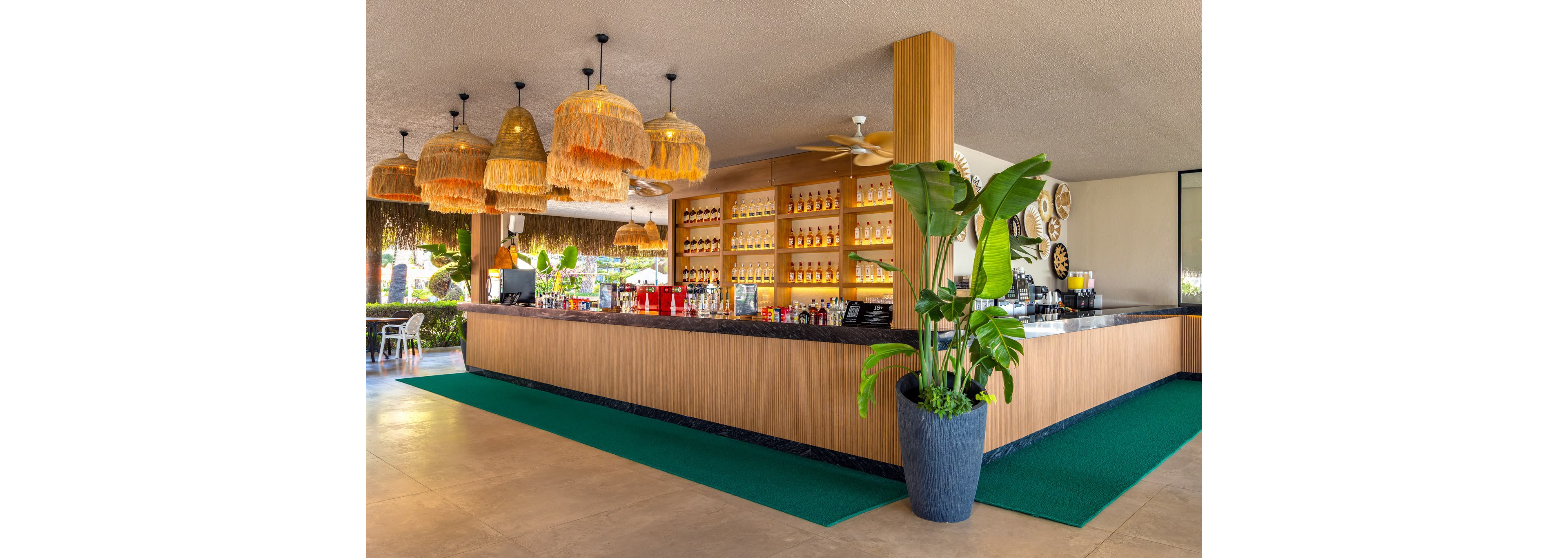 A contemporary open-air bar features a long counter with a neatly arranged wall of bottles behind it. Woven pendant lights hang from the ceiling, adding a warm, tropical touch to the space. A large potted plant stands beside the bar, complementing the natural wood finishes and green floor runners. The setting appears bright and inviting, suitable for relaxed social gatherings and vacation leisure.