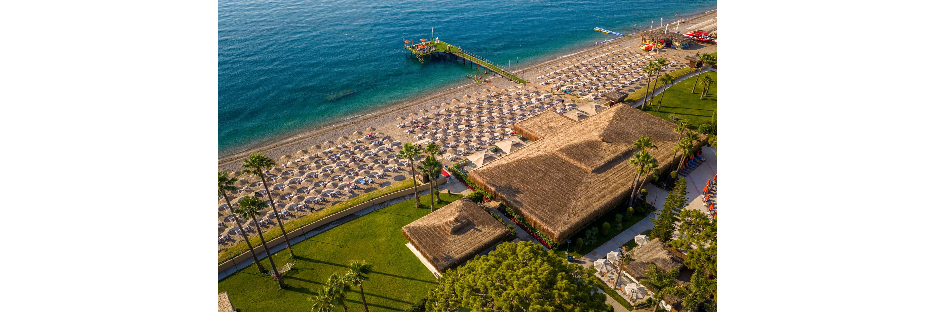 A large seaside resort is captured from above, showing rows of sun loungers and umbrellas lined up along a sandy beach. Calm blue sea water borders the shore, with a pier extending out and small structures at the end. In the foreground, thatched-roof buildings and manicured green lawns with palm trees frame the beachfront area. The scene suggests a popular vacation destination with organized facilities and a relaxed holiday atmosphere.