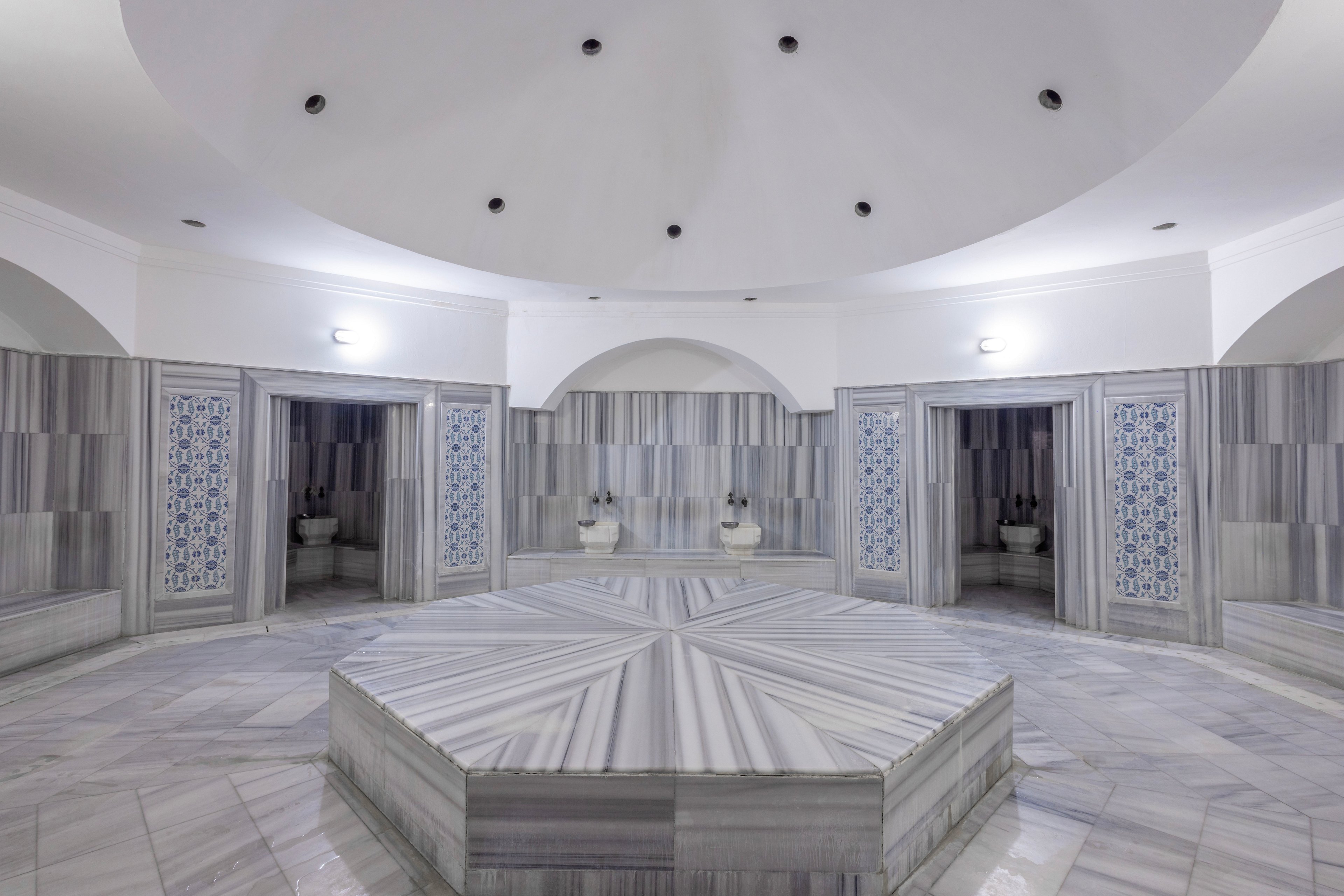 Symmetrical view of a traditional Turkish hammam with a large central marble platform. The room is surrounded by alcoves featuring marble basins and blue patterned tiles. Soft, even lighting highlights the white and gray striped marble surfaces and domed ceiling. The overall atmosphere is serene, clean, and spa-like, emphasizing relaxation and ritual bathing.