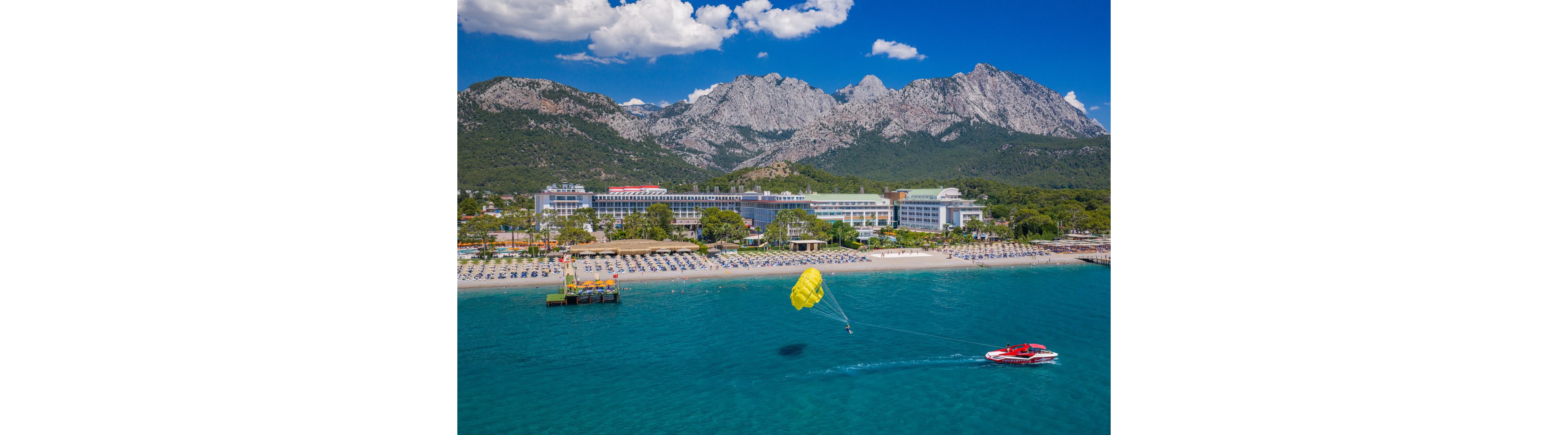 An aerial view shows a bright yellow parasail pulled by a red speedboat over clear turquoise water near a large beachfront resort. Rows of sun loungers line the sandy shore, with a pier extending into the sea. Lush green trees separate the hotel complex from dramatic rocky mountains rising in the background. The scene is bathed in sunny, clear weather with scattered white clouds in the blue sky.