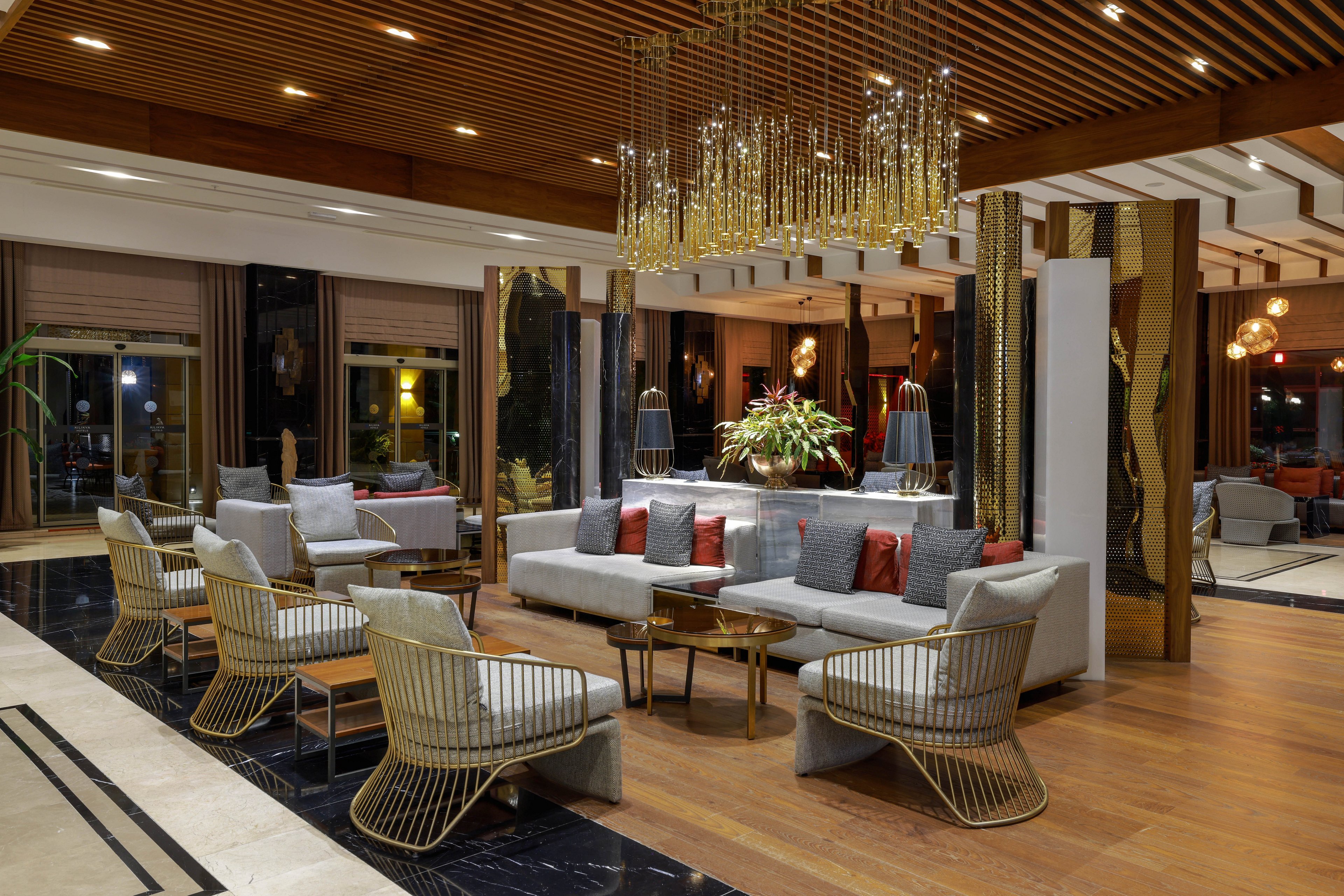 A contemporary hotel lobby lounge features a central seating area with a long sofa and multiple armchairs arranged around low coffee tables. Warm wooden flooring and a slatted wood ceiling create a cozy yet upscale atmosphere. A sculptural chandelier with hanging metallic elements adds visual interest above the seating. Large windows and glass doors reveal the nighttime exterior, while indoor plants and decorative panels enhance the stylish interior design.