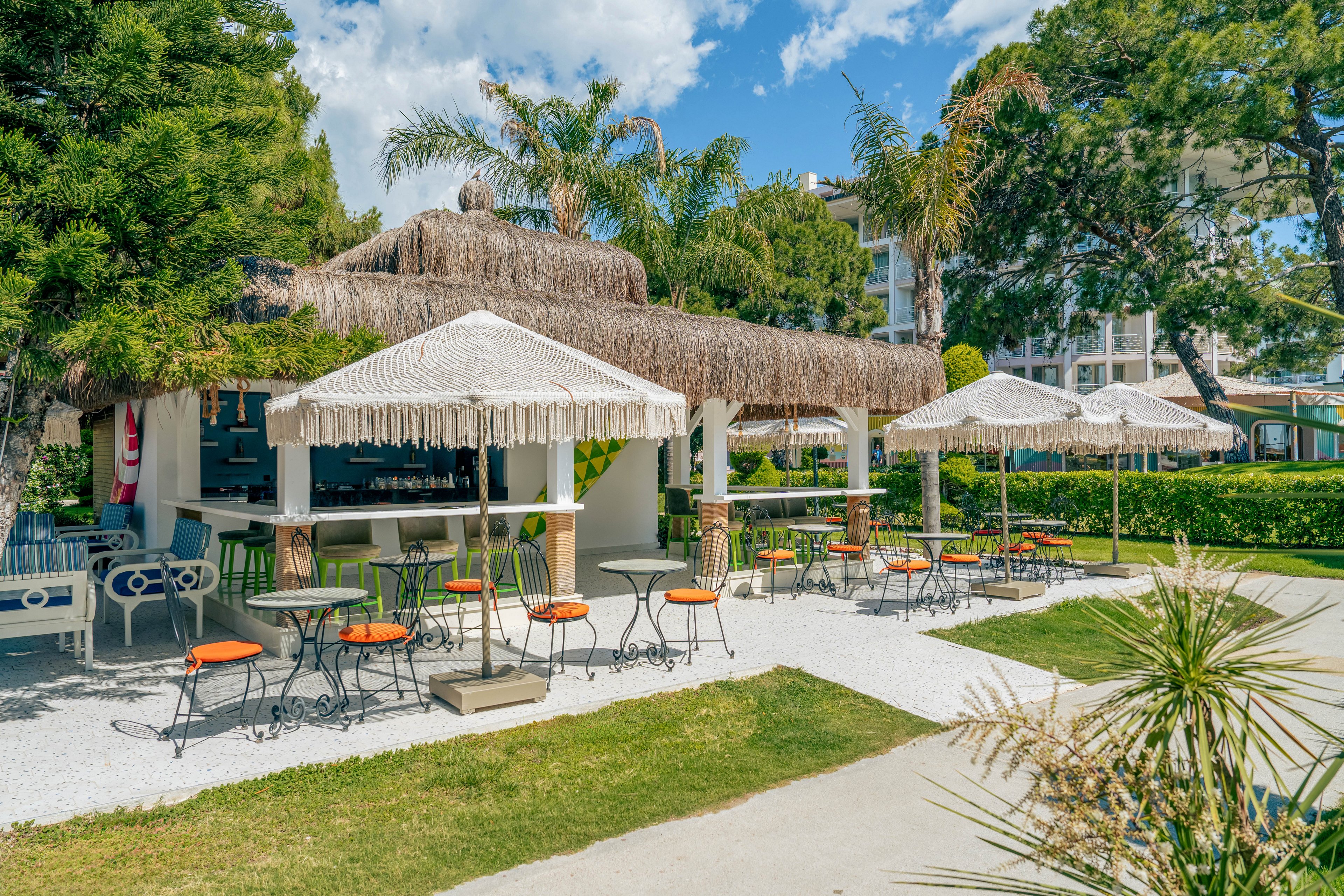 An open-air bar with a thatched roof is set in a lush garden under a bright blue sky. Round tables with orange-seated chairs are arranged on a gravel patio, offering shaded seating beneath straw umbrellas. Palm trees and green shrubs surround the area, creating a relaxed resort atmosphere. In the background, modern buildings and manicured lawns suggest a hotel or vacation property.