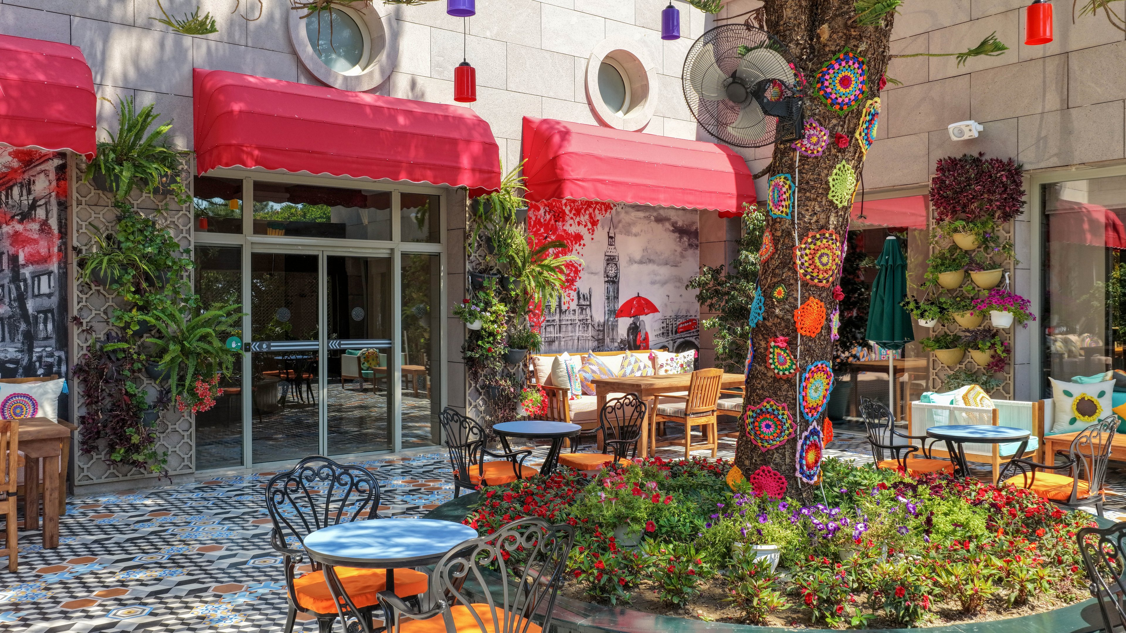 An inviting outdoor café features colorful chairs and small round tables arranged around a central tree. The tree is wrapped in bright crochet decorations and surrounded by a lush flower bed. Red awnings shade the building façade, which is accented with hanging plants and decorative wall art. Multicolored lanterns hang above the patio, creating a cheerful and artistic atmosphere. The scene suggests a relaxed, cozy spot for dining or coffee in a lively urban courtyard.