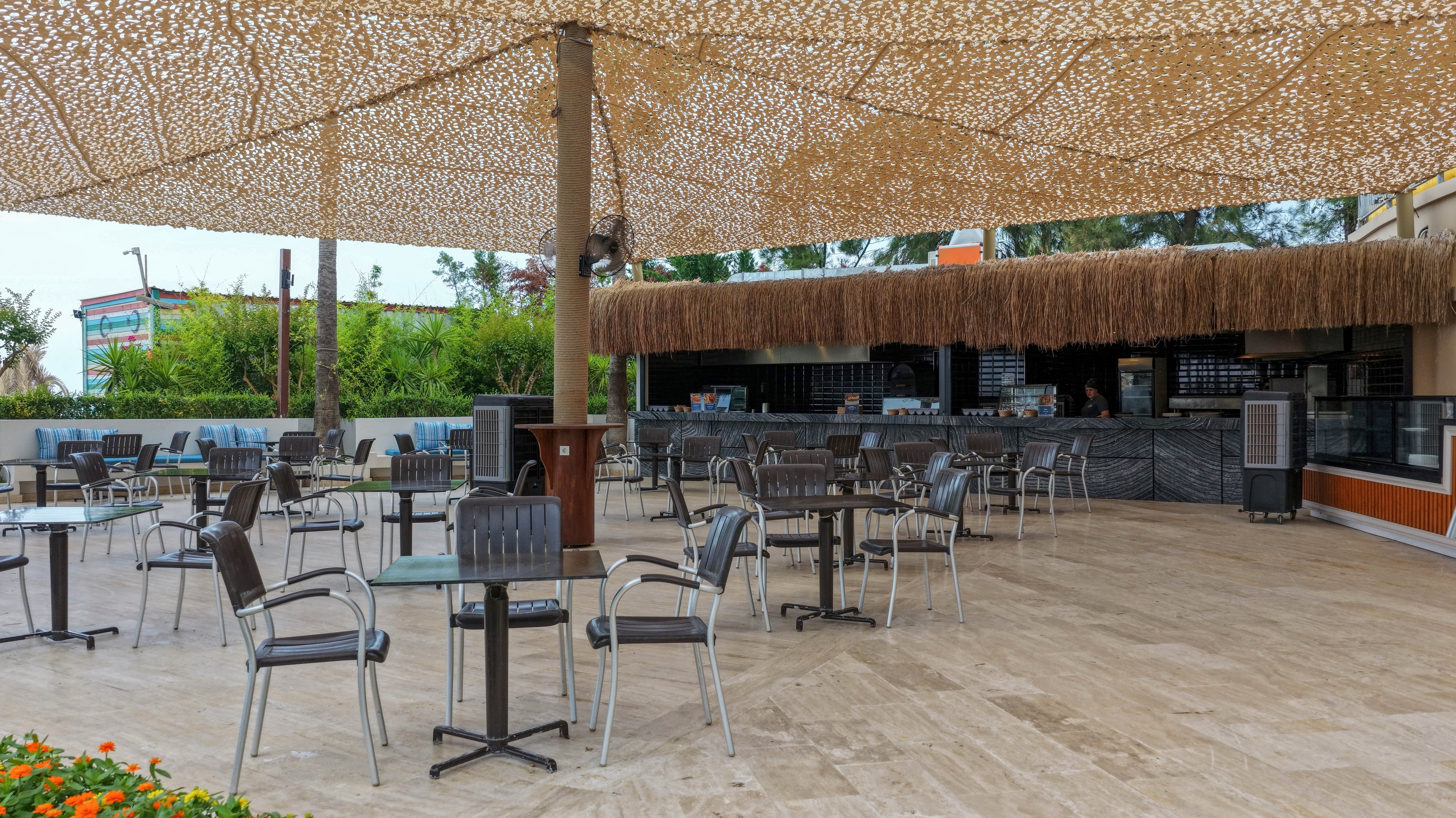 An open-air cafe terrace features multiple empty tables and chairs arranged on a spacious stone floor. A large woven canopy provides shade over the seating area, creating a patterned light effect. The bar counter in the background has a thatched roof detail and modern finishes. Greenery and flowers border the space, giving the outdoor setting a relaxed, resort-like atmosphere.