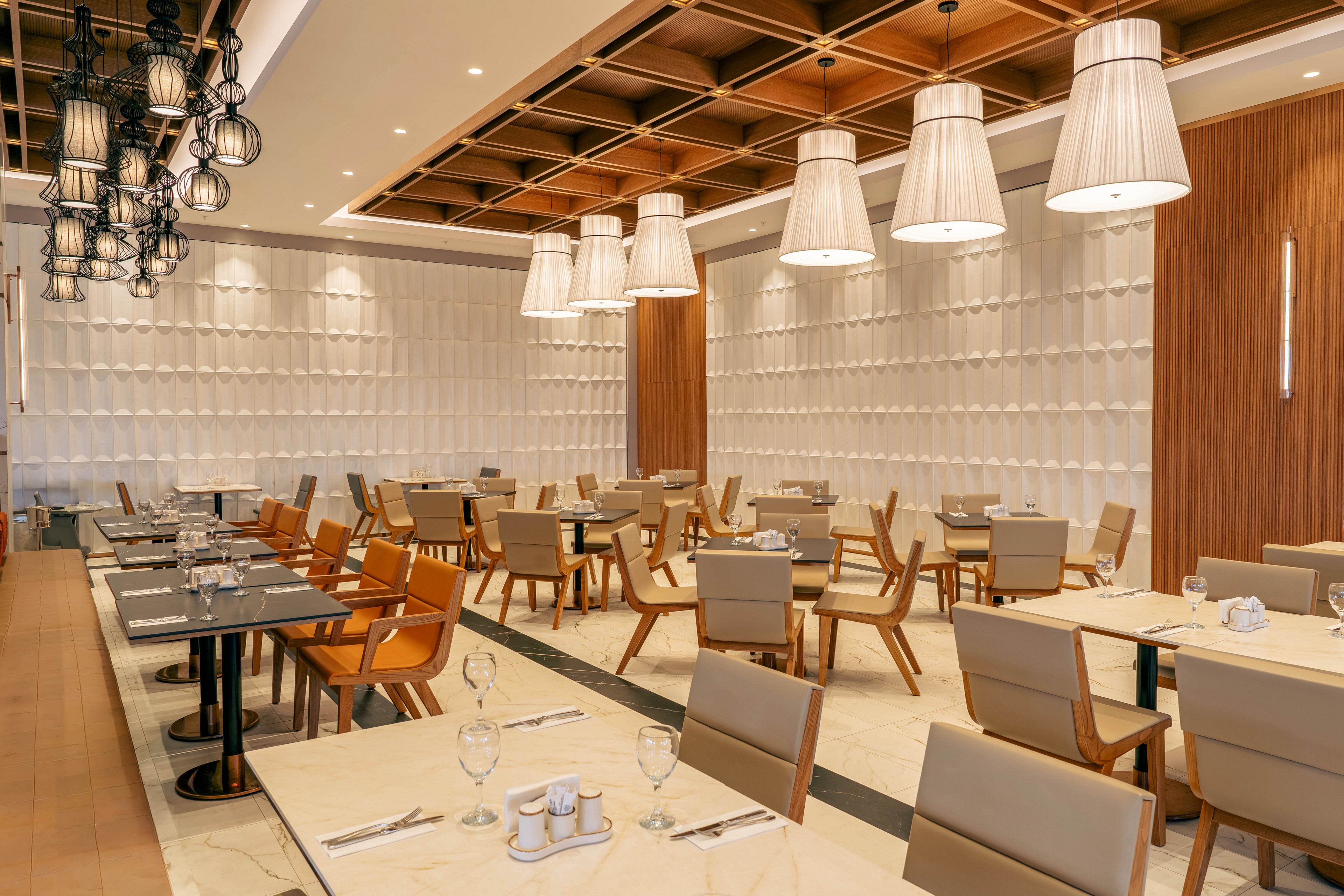 A contemporary restaurant dining area is shown with neatly arranged tables and chairs. The space features a patterned white wall, wooden accents, and a decorative ceiling with large pendant lights. Tables are set with glassware, plates, and condiments, suggesting readiness for guests. The overall atmosphere is clean, organized, and warmly lit, creating an inviting yet quiet setting.