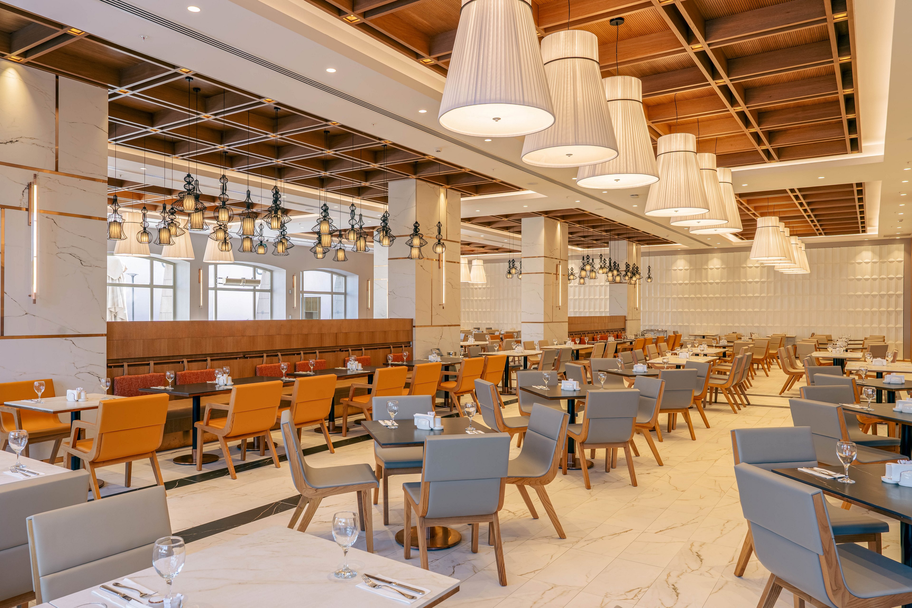 A spacious modern restaurant dining area is shown with neatly arranged tables and chairs. The room features a warm neutral color palette with orange and gray seating and light marble flooring. Large pendant lights hang from a wooden ceiling grid, creating a cozy yet upscale ambiance. Table settings with glassware and cutlery are prepared, but no guests are present, emphasizing a clean and orderly environment.